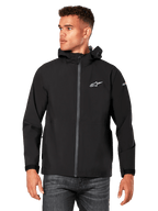 Male model wearing Alpinestars Kascade Rain Jacket, Black, white chest logo and sleeve branding, hooded rain shell with coated front zipper and velcro cuffs, paired with dark jeans, casual motorsport-inspired streetwear