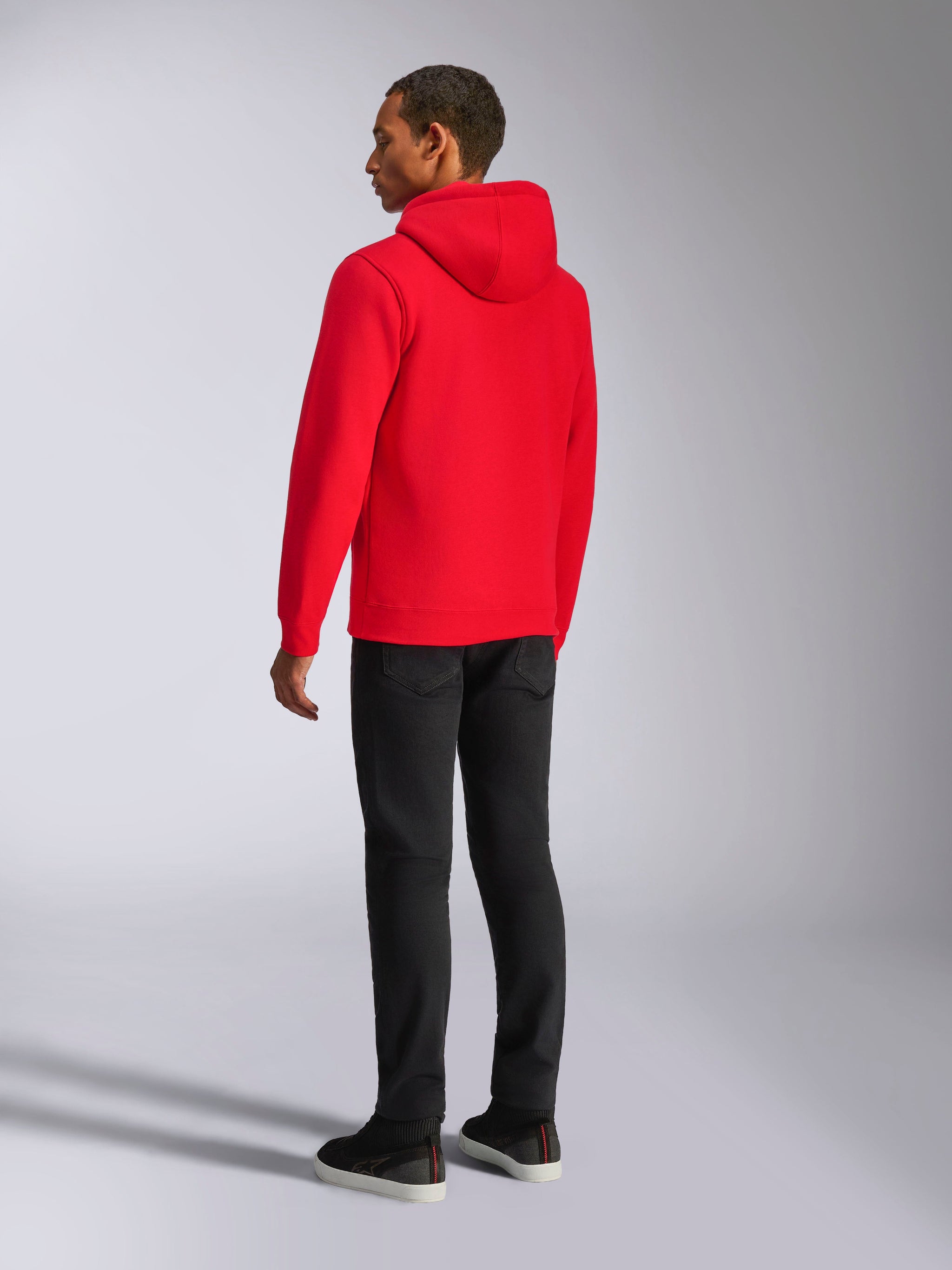 Alpinestars Ageless V3 Hoodie, casual streetwear, vibrant red, rear view of model, featuring jersey-lined hood, ribbed cuffs and hem, paired with black pants, modern motorsport-inspired design.