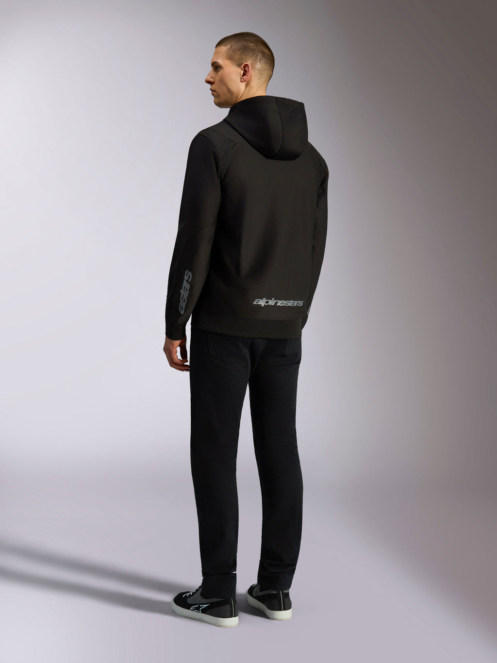 Alpinestars Sessions V3 Hoodie, black casual streetwear, back view on a model, featuring grey alpinestars wordmark on the lower back and Astars logo on the sleeve, hooded zip-front design with modern motorsport-inspired styling