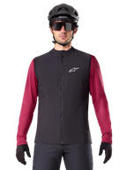 Alpinestars A-Dura Thermal Vest, Black insulated cycling vest, worn by a model with a maroon long-sleeve jersey, black MTB helmet, and sunglasses, featuring a white logo on the chest, lightweight thermal protection for mountain biking