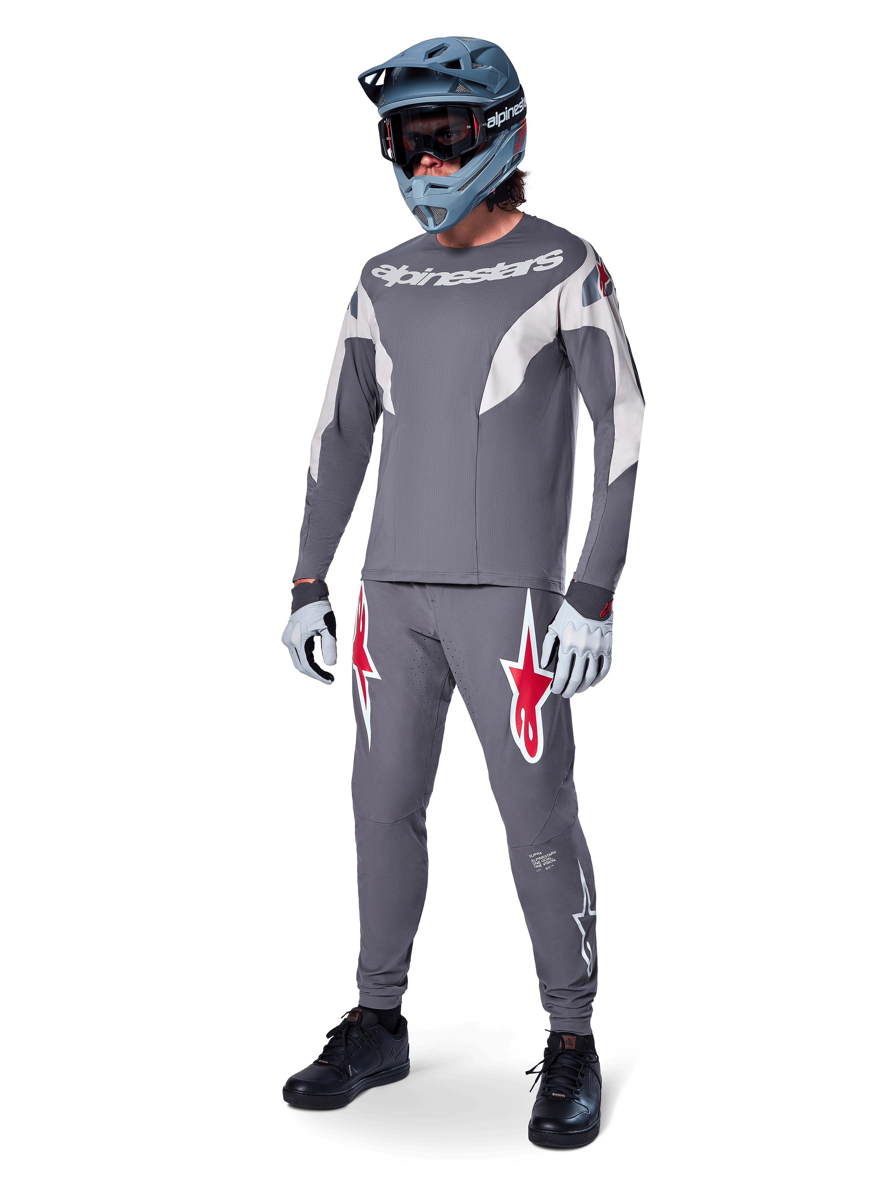 Alpinestars A-Supra Race Pants in Dark Gray, featuring bold red and white logos on the thighs, worn by a rider as part of a complete mountain bike racing kit with a matching gray jersey, blue-gray full-face helmet, and light gray gloves, high-performance gear for MTB racing