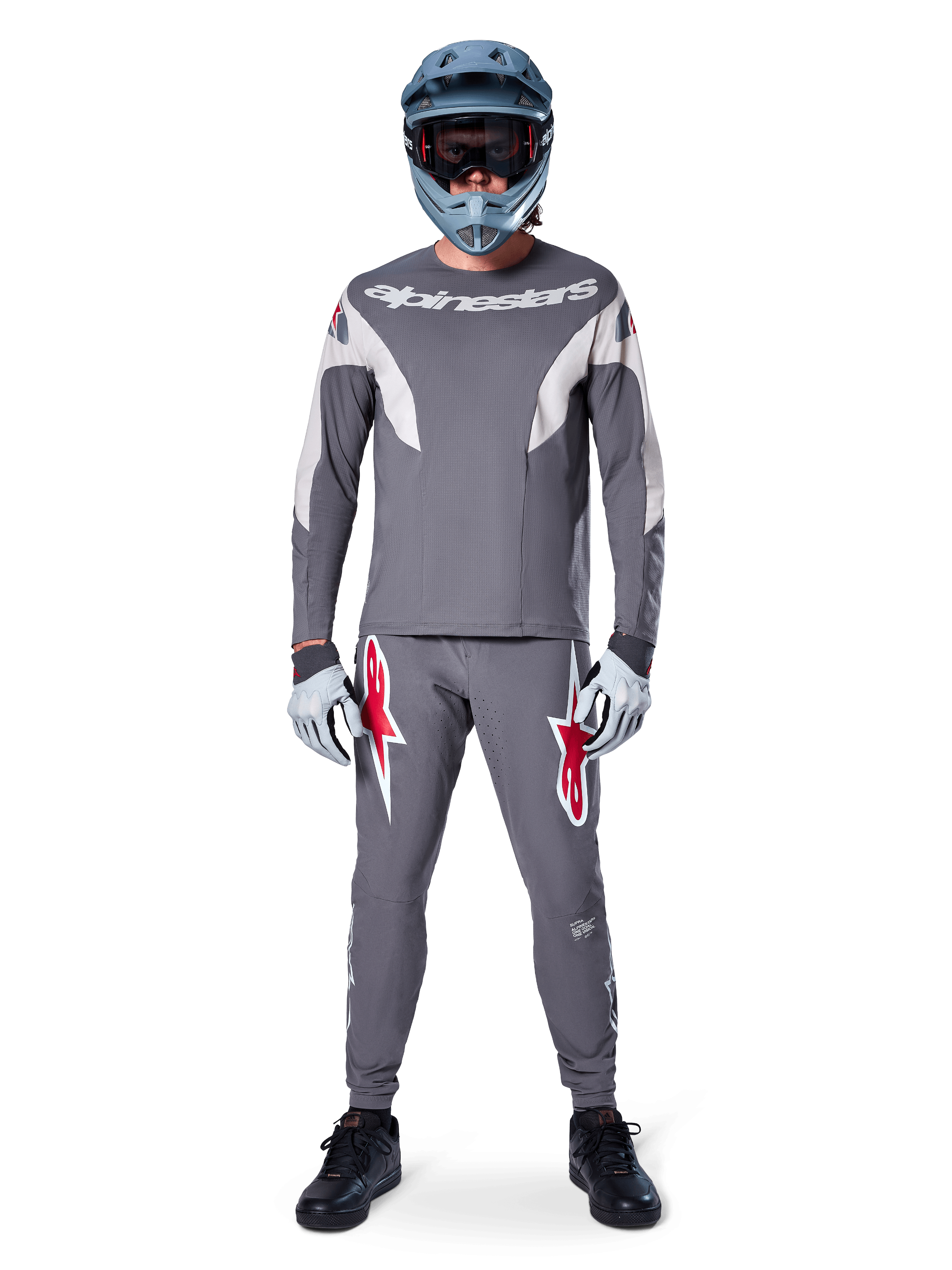 MTB rider wearing Alpinestars A-Supra Race Pants in Dark Gray, featuring bold red logo accents on the thighs, paired with a matching gray jersey, blue full-face helmet, and black shoes for a complete off-road racing kit