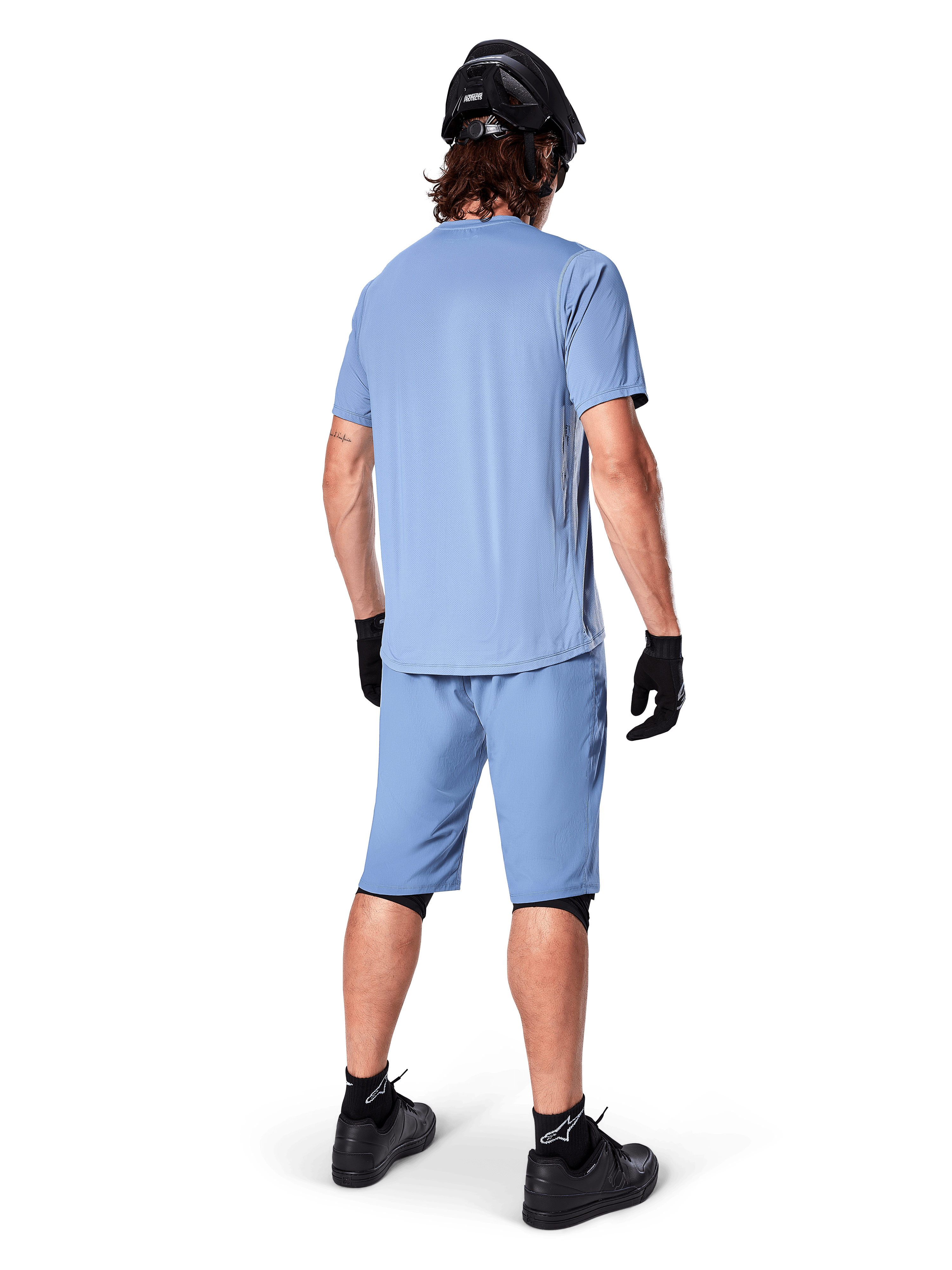 Rear view of a mountain biker wearing Alpinestars A-Dura Shorts in Infinity Blue, matching short-sleeve jersey, black helmet, black gloves, and black shoes, coordinated performance gear for MTB trail riding