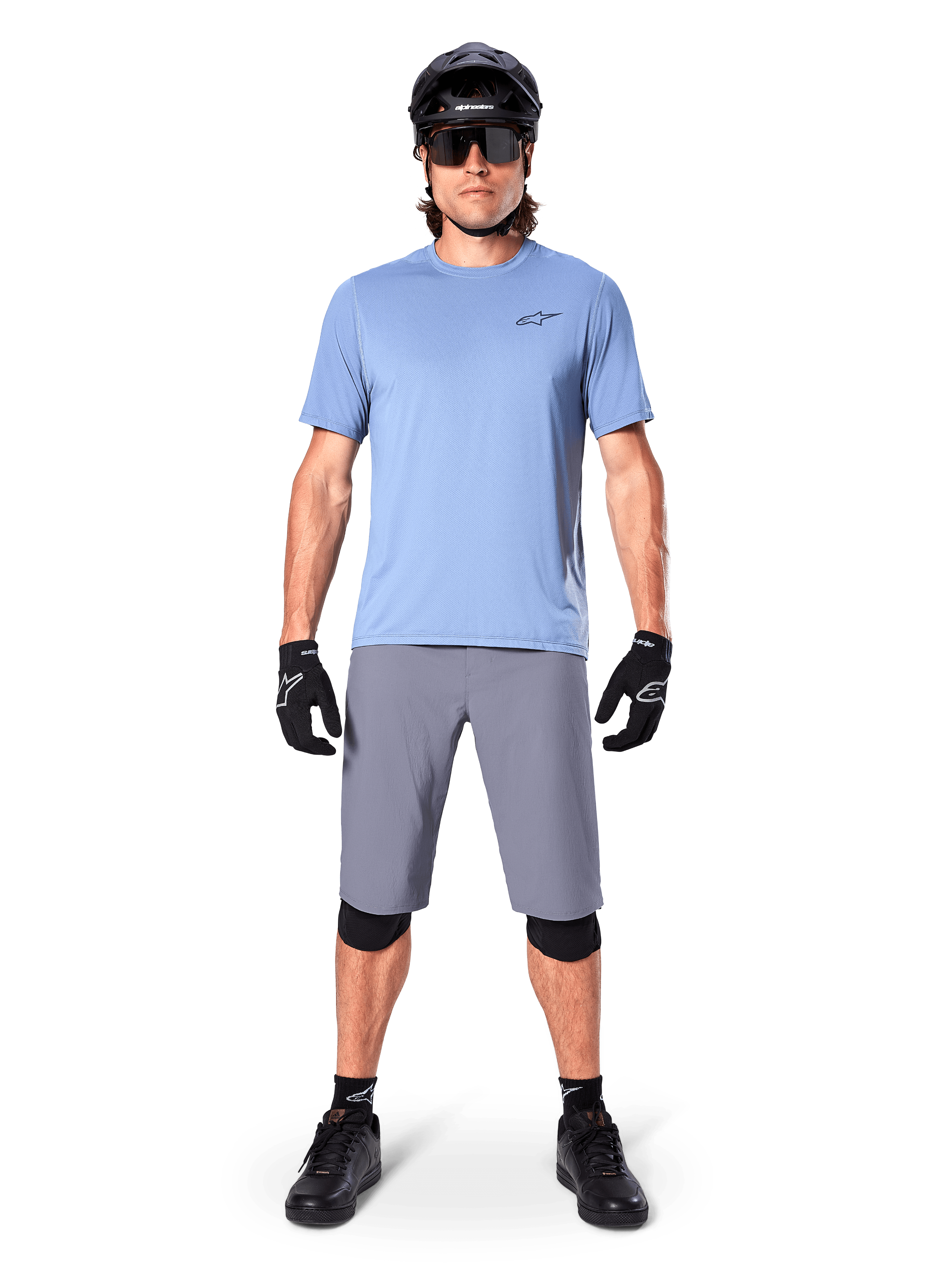 Male rider wearing Alpinestars A-Dura Shorts in Dark Gray, styled with a light blue short-sleeve t-shirt, black helmet, sunglasses, and black knee pads, complete mountain biking outfit for off-road performance.