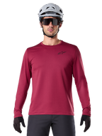 Male cyclist wearing Alpinestars A-Dura Thermal Jersey, Long Sleeve MTB Jersey, Burgundy, styled with a gray helmet and black sunglasses, features a small black star logo on the chest, insulated fleece fabric for cold-weather mountain biking