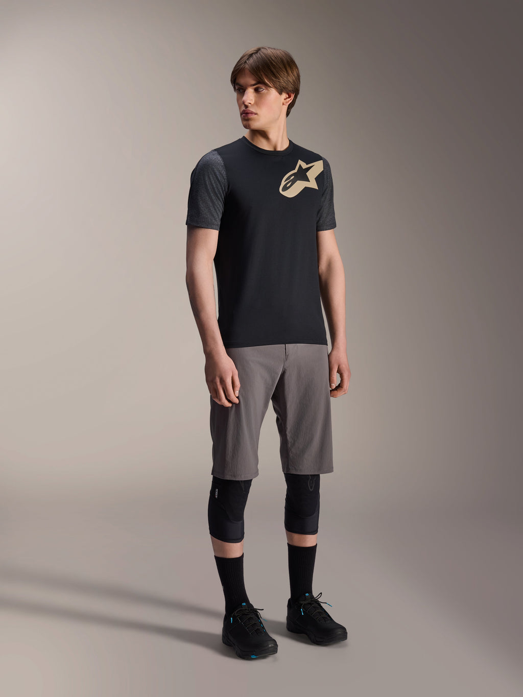 Male model wearing Alpinestars A-Dura Lipan Short Sleeve Jersey in Black, featuring dark grey sleeves and a tan Astars logo on the chest, paired with grey mountain bike shorts, black knee pads, and black cycling shoes, coordinated mountain bike riding gear