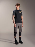 Male model wearing Alpinestars A-Dura Lipan Short Sleeve Jersey in Black, featuring dark grey sleeves and a tan Astars logo on the chest, paired with grey mountain bike shorts, black knee pads, and black cycling shoes, coordinated mountain bike riding gear