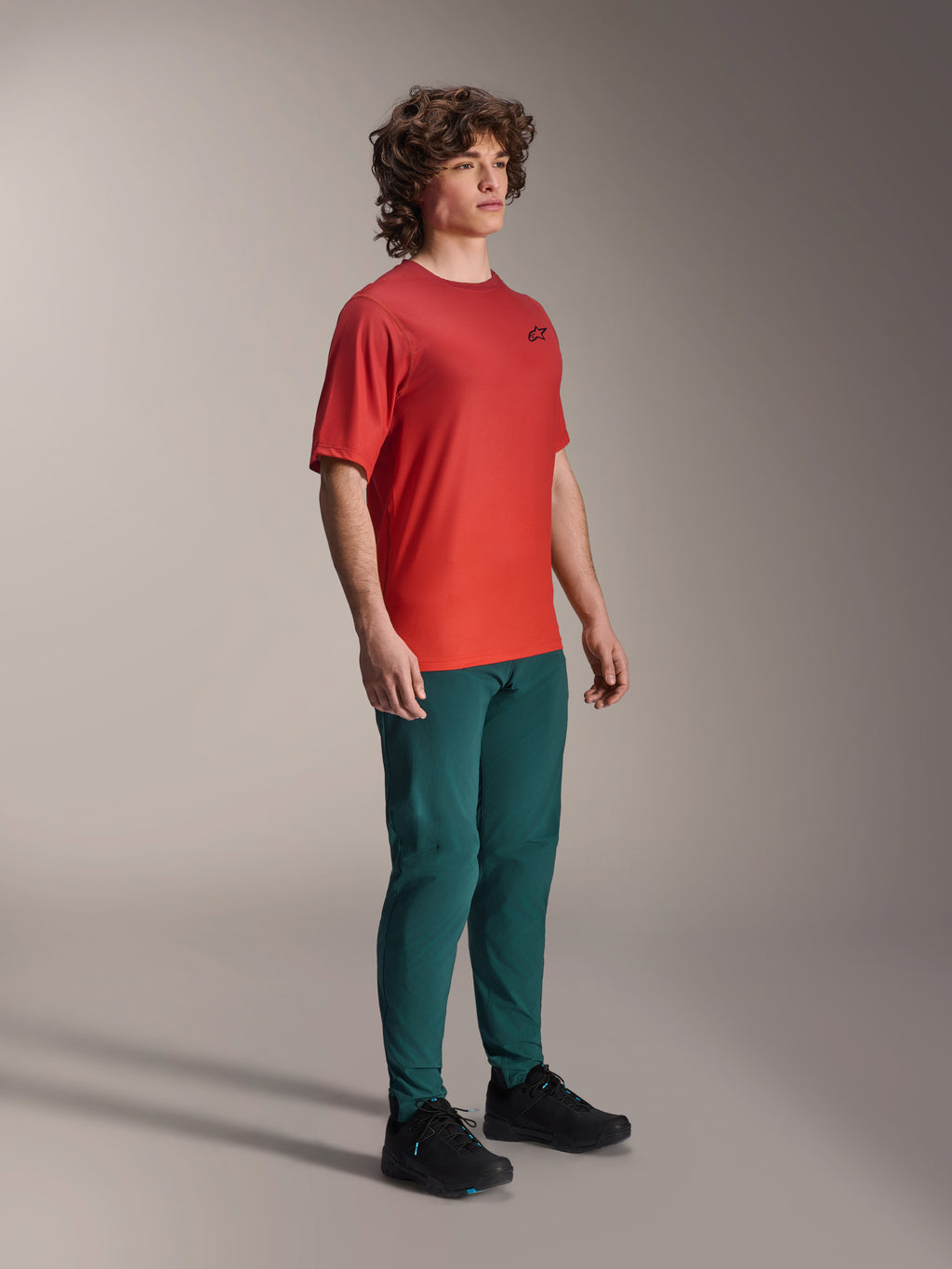 Model wearing Alpinestars A-Dura Pants in Dark Pine, paired with a red A-Dura Rise Short Sleeve Jersey and black MTB shoes, athletic mountain bike apparel in a studio setting