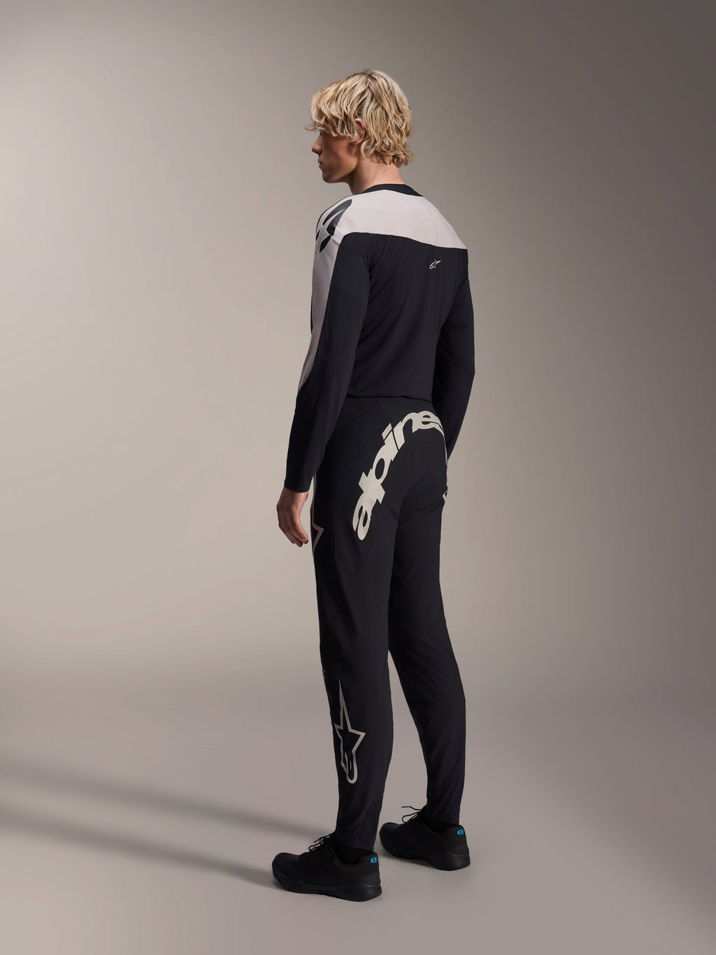 Alpinestars A-Supra Melt Pants, MTB Pants, Black, featuring a large white Alpinestars wordmark on the back yoke and star logo on the leg, worn by a model with a matching long-sleeve jersey and black shoes, back-side view