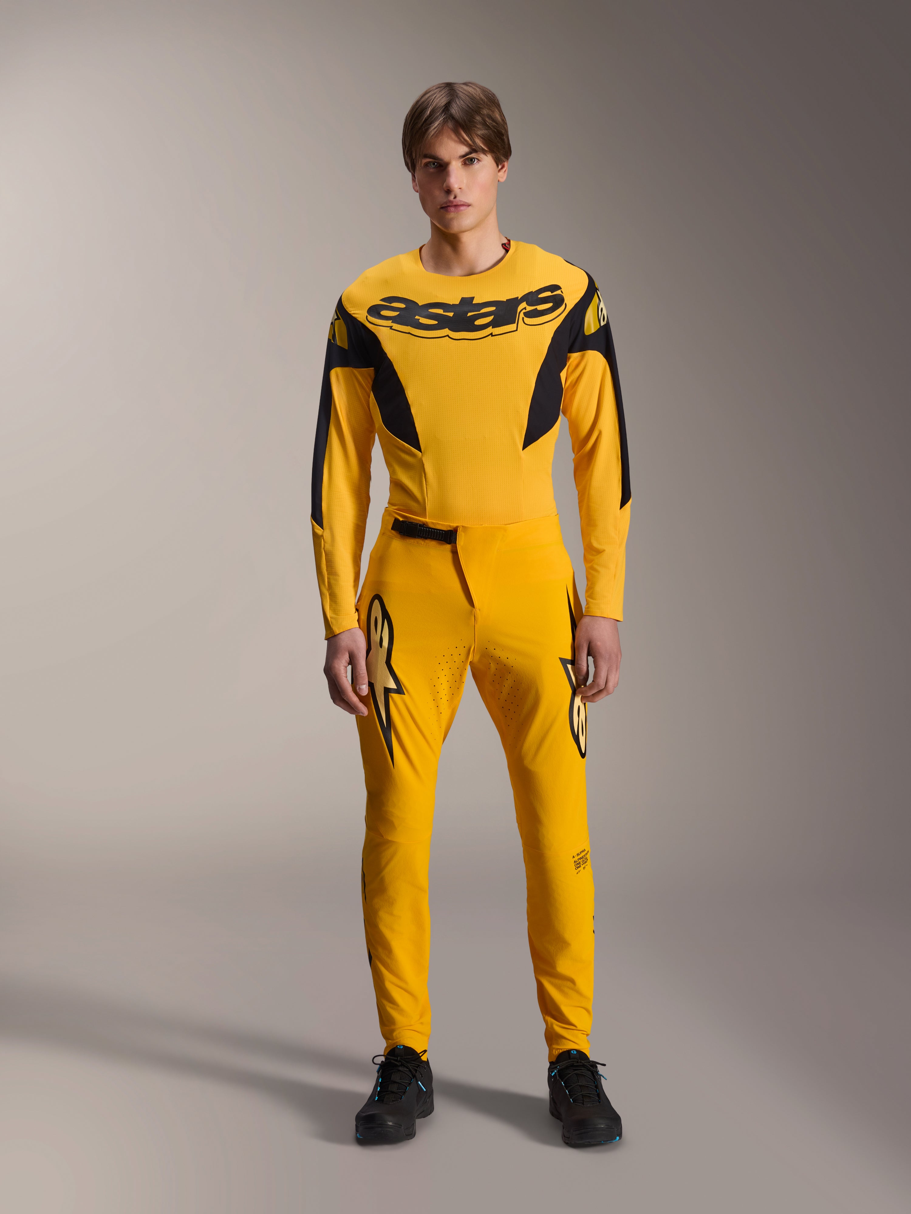 Limited Edition A-Supra Melt Gold Gear Set, Alpinestars Mountain Bike Apparel, Dark Yellow and Black, Model wearing long-sleeve jersey with bold 'astars' chest logo and matching performance pants, featuring laser-cut ventilation on inner thighs and a secure ratchet waist closure, professional MTB racing kit