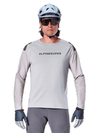 Alpinestars A-Aria Polartec Switch Long Sleeve Jersey, Light Gray mountain bike jersey featuring laser perforations on sleeves and chest branding, worn with light blue helmet, sunglasses, and gray gloves