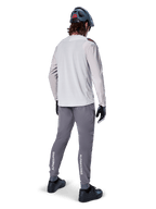Rear view of a cyclist wearing the Alpinestars A-Aria Polartec Switch Long Sleeve Jersey in Light Gray, paired with dark gray mountain bike pants featuring white Alpinestars branding on the calves, black gloves, and a gray helmet.