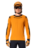Male model wearing Alpinestars A-Dura Dri Wool Long Sleeve Jersey in Dark Gold, coordinated with a matching gold MTB helmet and gloves, black mesh panels on shoulders and sleeves, black Alpinestars wordmark on chest, dark sunglasses and pants, technical mountain bike apparel
