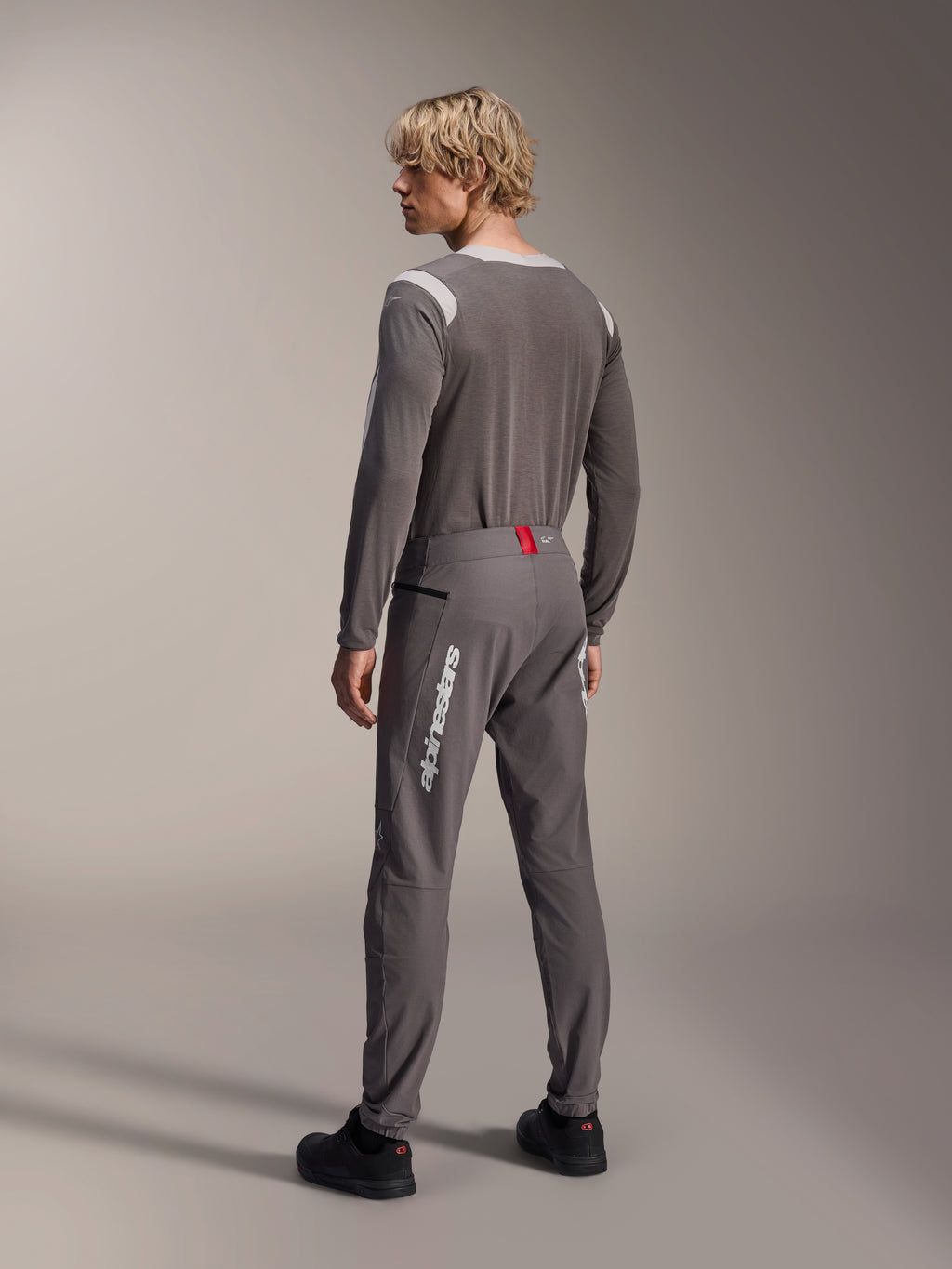 Model from a rear view wearing Alpinestars MTB gear, gray A-Dura Dri Wool Long Sleeve Jersey, Dark Gray A-Dura Elite Pants with white vertical branding and red waistband detail, coordinated cycling outfit for mountain biking