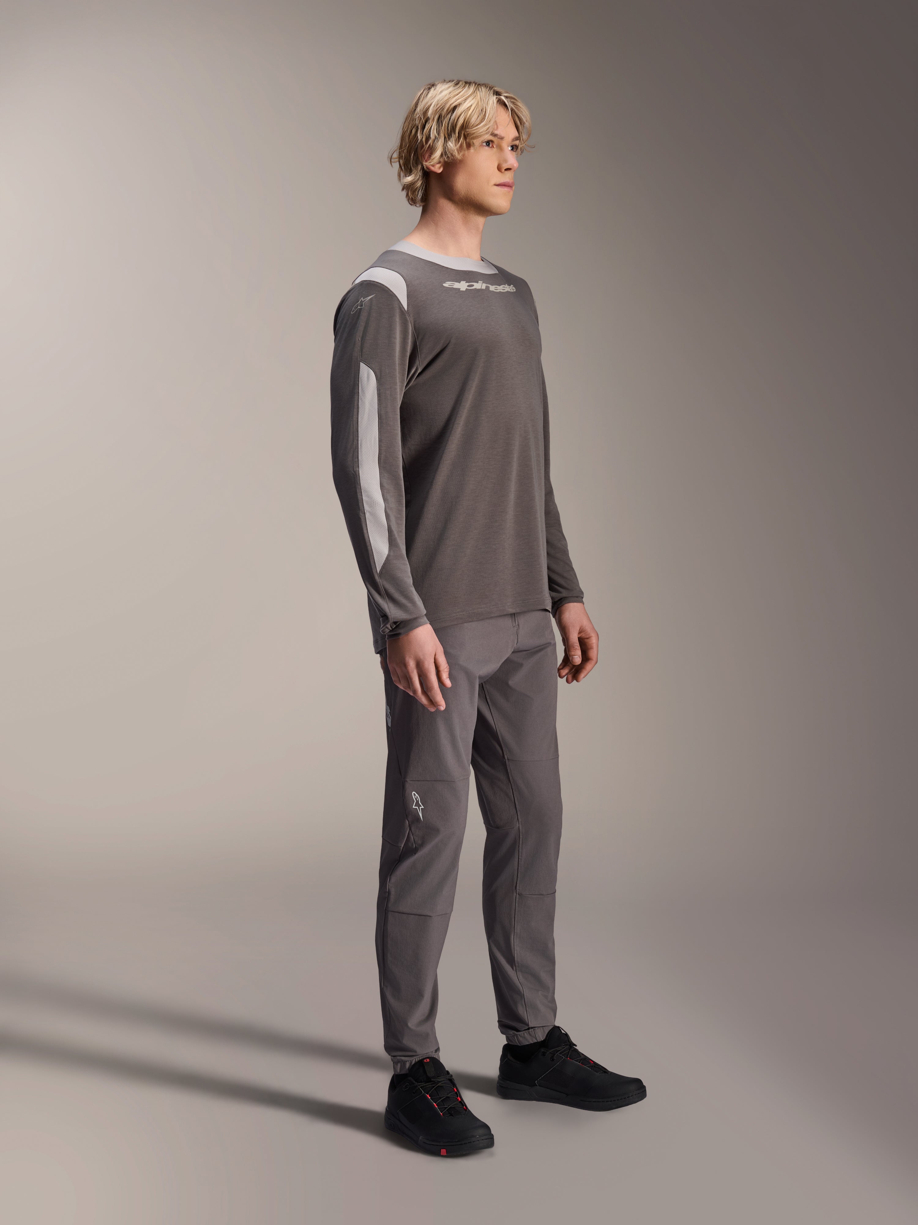 Male model wearing Alpinestars A-Dura Dri Wool Long Sleeve Jersey in Dark Gray, paired with matching gray pants and black shoes, featuring light gray mesh panels on the shoulders and sleeves, technical MTB gear for off-road cycling