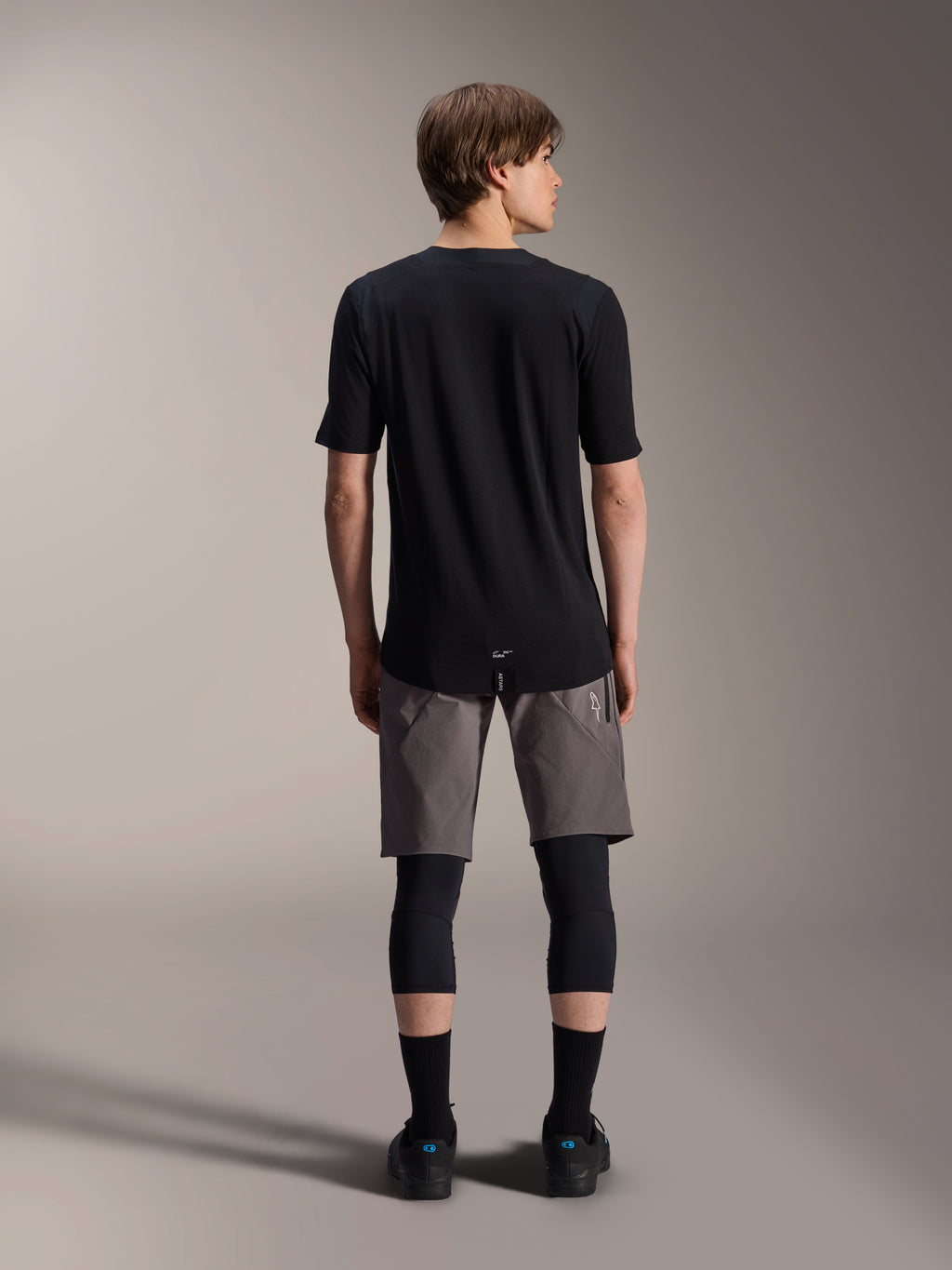 Alpinestars A-Dura Dri Wool Short Sleeve Jersey, Black, back view worn by model with grey MTB shorts and black knee guards, featuring a longer drop tail and athletic fit for off-road cycling