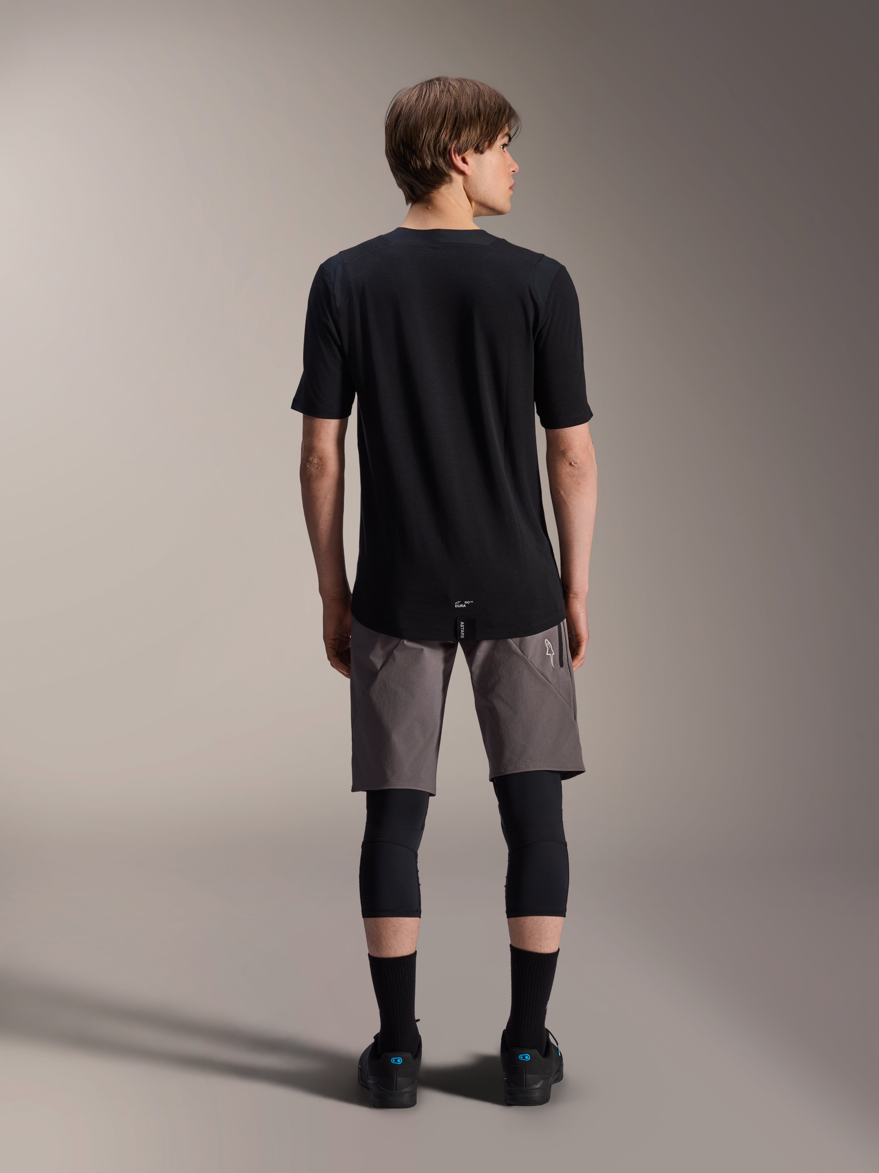 Alpinestars A-Dura Dri Wool Short Sleeve Jersey, Black, back view worn by model with grey MTB shorts and black knee guards, featuring a longer drop tail and athletic fit for off-road cycling