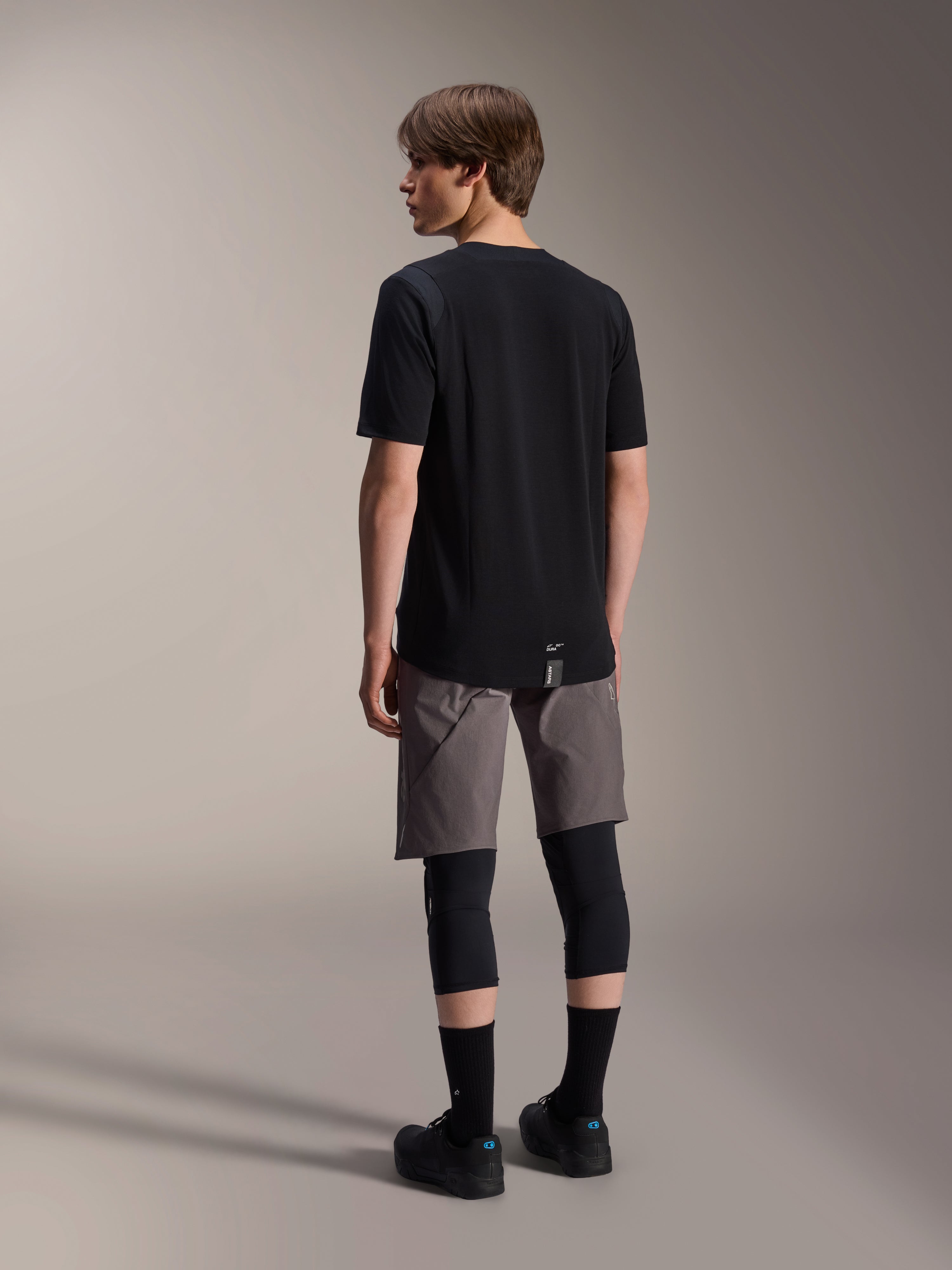 Alpinestars A-Dura Dri Wool Short Sleeve Jersey, Black, back view on model, worn with grey mountain biking shorts and black knee guards, featuring a dropped tail hem and subtle logo, technical MTB apparel