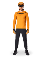 Alpinestars A-Dura Rocker Long Sleeve Jersey in Dark Gold, featuring a black screen-printed logo on the chest, worn with a matching gold MTB helmet, black sunglasses, and black pants, professional mountain biking gear