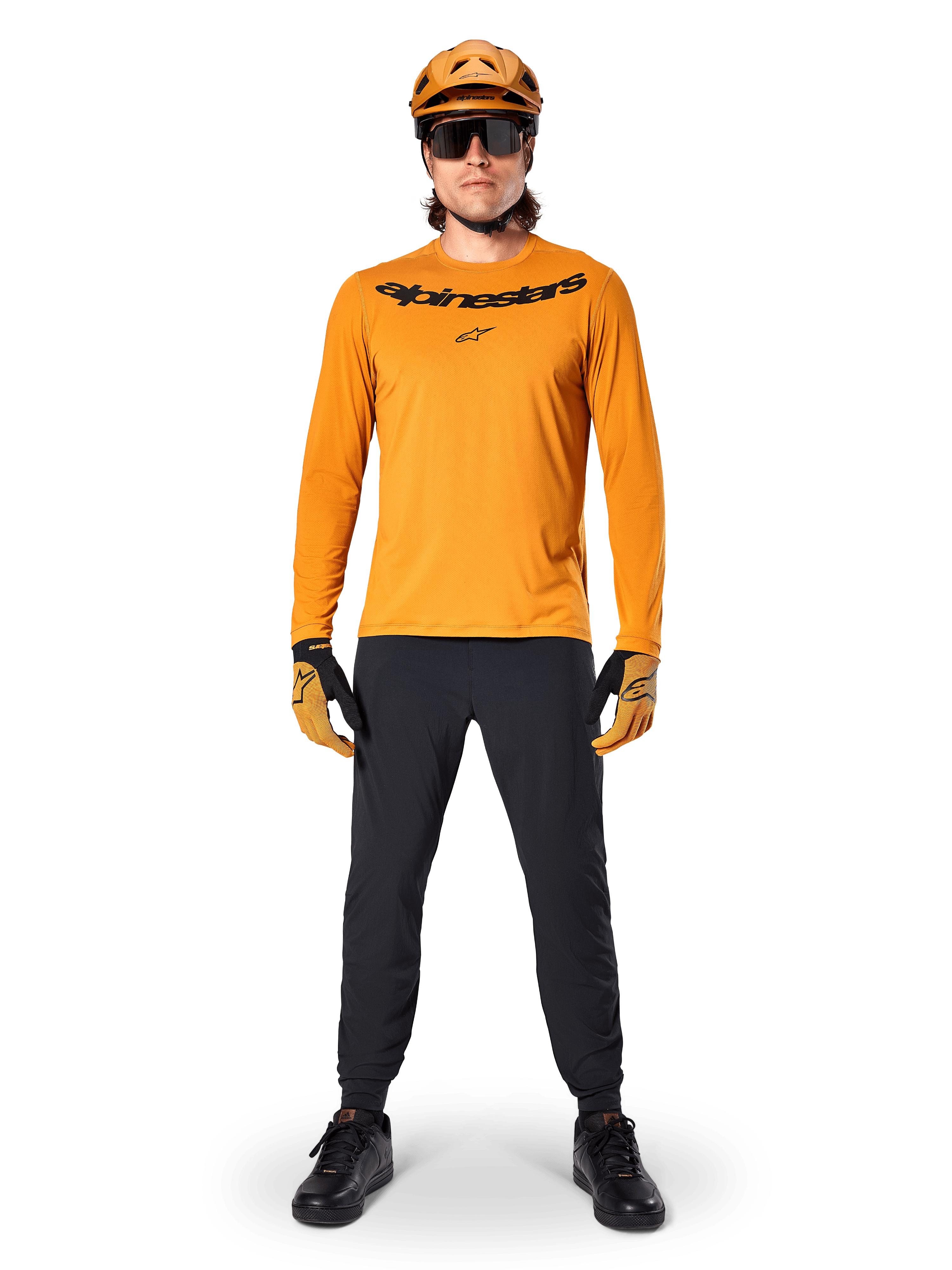 Alpinestars A-Dura Rocker Long Sleeve Jersey in Dark Gold, featuring a black screen-printed logo on the chest, worn with a matching gold MTB helmet, black sunglasses, and black pants, professional mountain biking gear