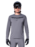 Male rider wearing the Alpinestars A-Dura Rocker Long Sleeve Jersey, Dark Gray MTB jersey with black wordmark on chest, paired with gray pants, black helmet, and gloves, designed for off-road cycling