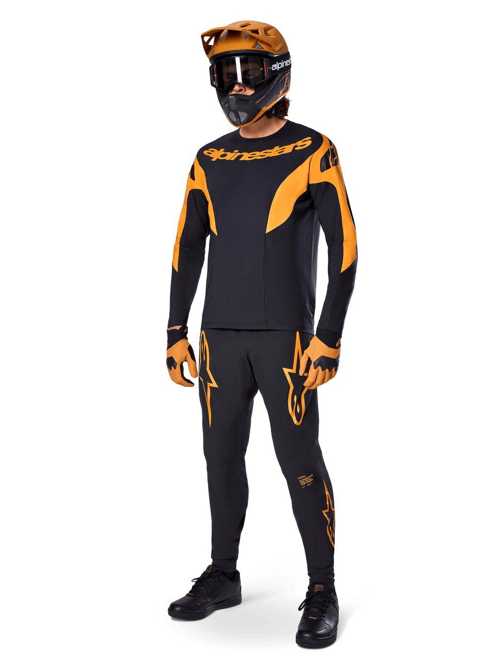 MTB rider wearing complete racing gear, Black A-Supra Race Jersey with orange accents, matching black pants with orange Alpinestars logos, orange and black full-face helmet with goggles, orange and black gloves, coordinated outfit for mountain bike racing
