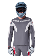 MTB rider wearing Alpinestars A-Supra Race Jersey - Long Sleeve in Dark Gray, long-sleeve cycling jersey with white Alpinestars branding, paired with a blue-gray full-face helmet, goggles, and gray racing gloves, complete mountain bike racing gear for off-road performance