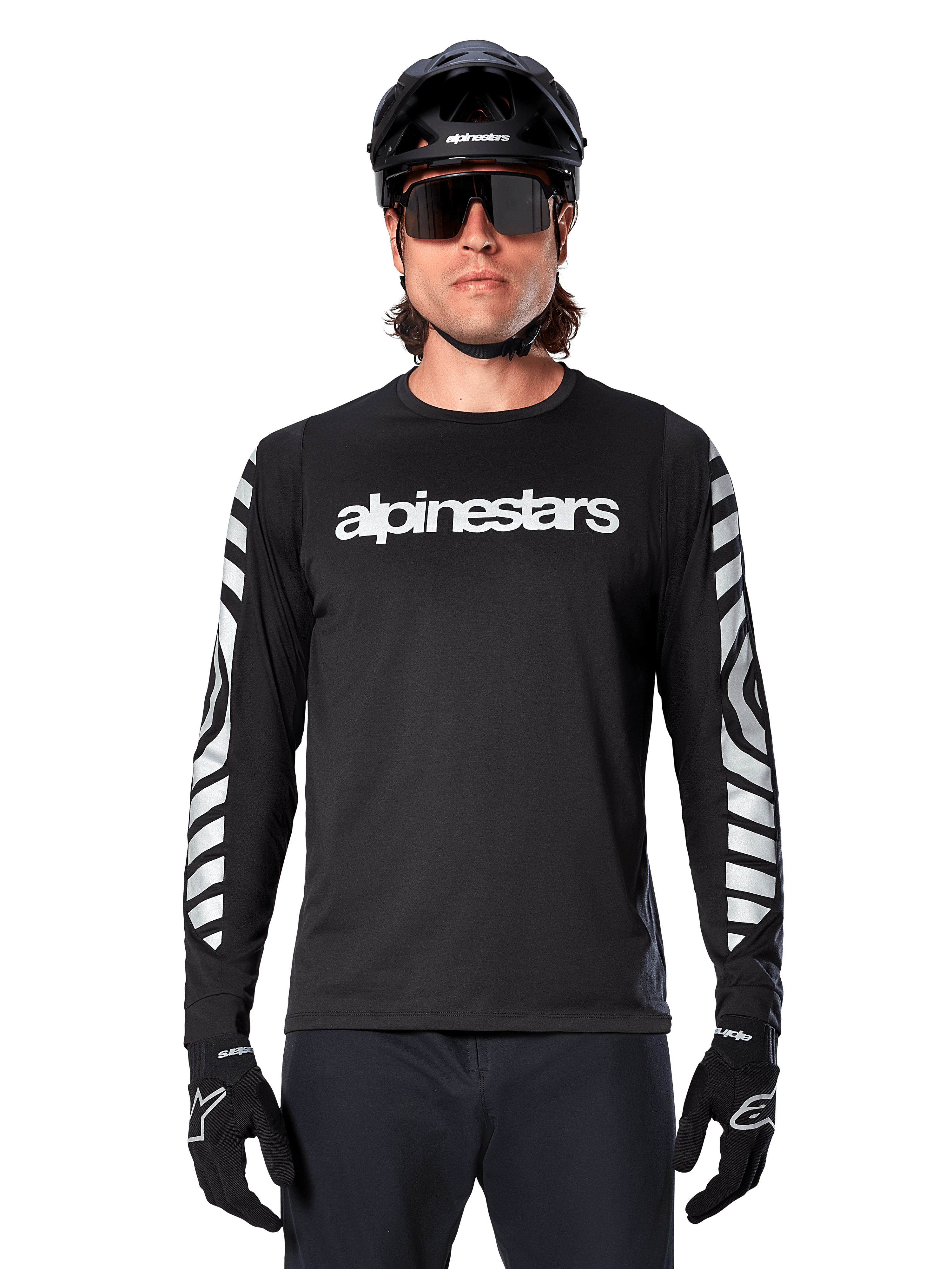 Male rider wearing Alpinestars A-Dura Dri Oscar Long Sleeve Jersey, Black Reflective, featuring white alpinestars wordmark on chest and silver reflective graphics on sleeves, paired with black MTB helmet, sunglasses, and black gloves, performance cycling gear