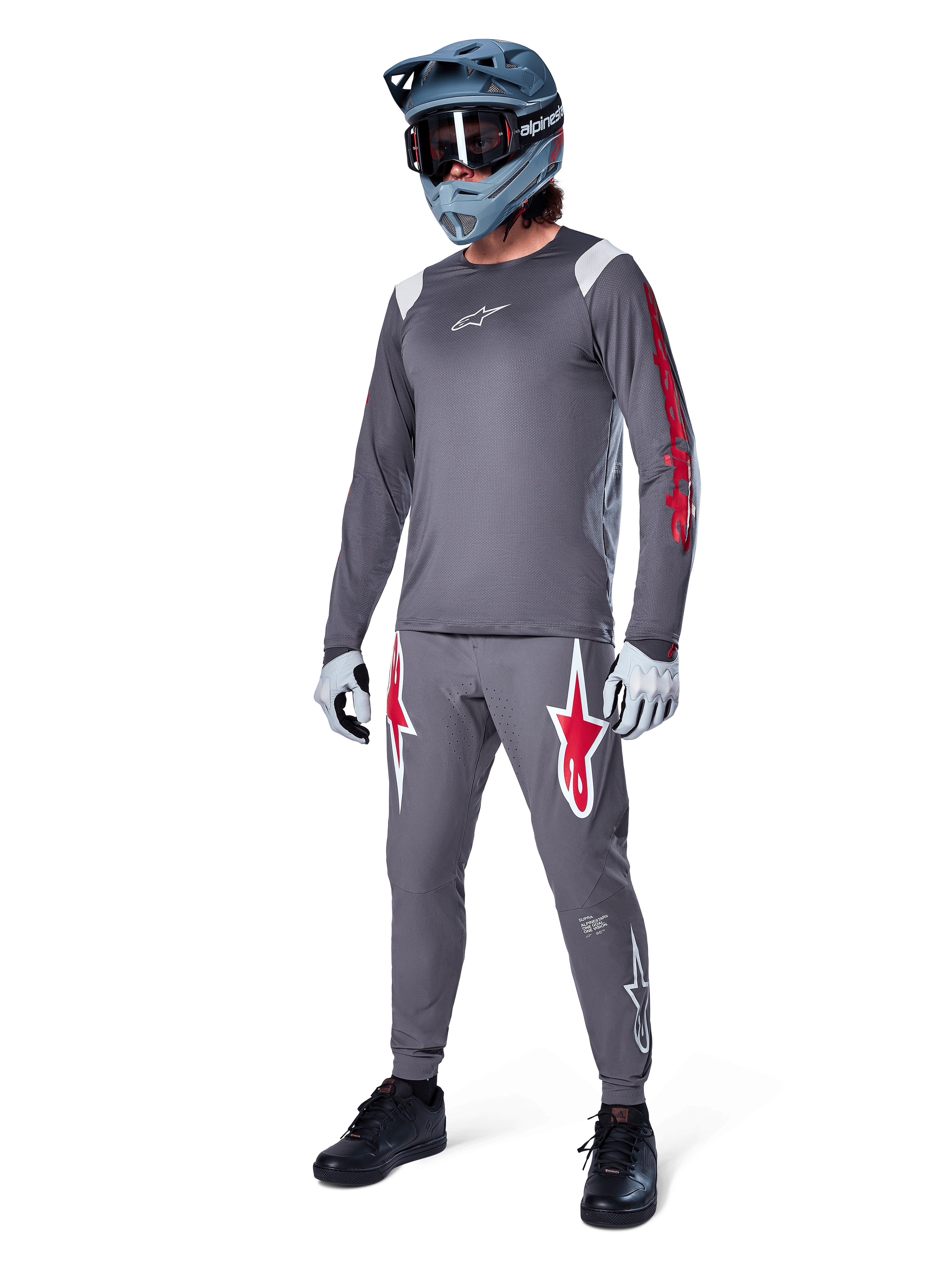 Alpinestars A-Supra Astar Long Sleeve Jersey in Dark Gray, featuring red branding on the sleeves and a white logo on the chest, worn by a rider with matching gray pants, a blue-gray full-face helmet, and performance gloves, coordinated mountain bike gear for gravity riding.