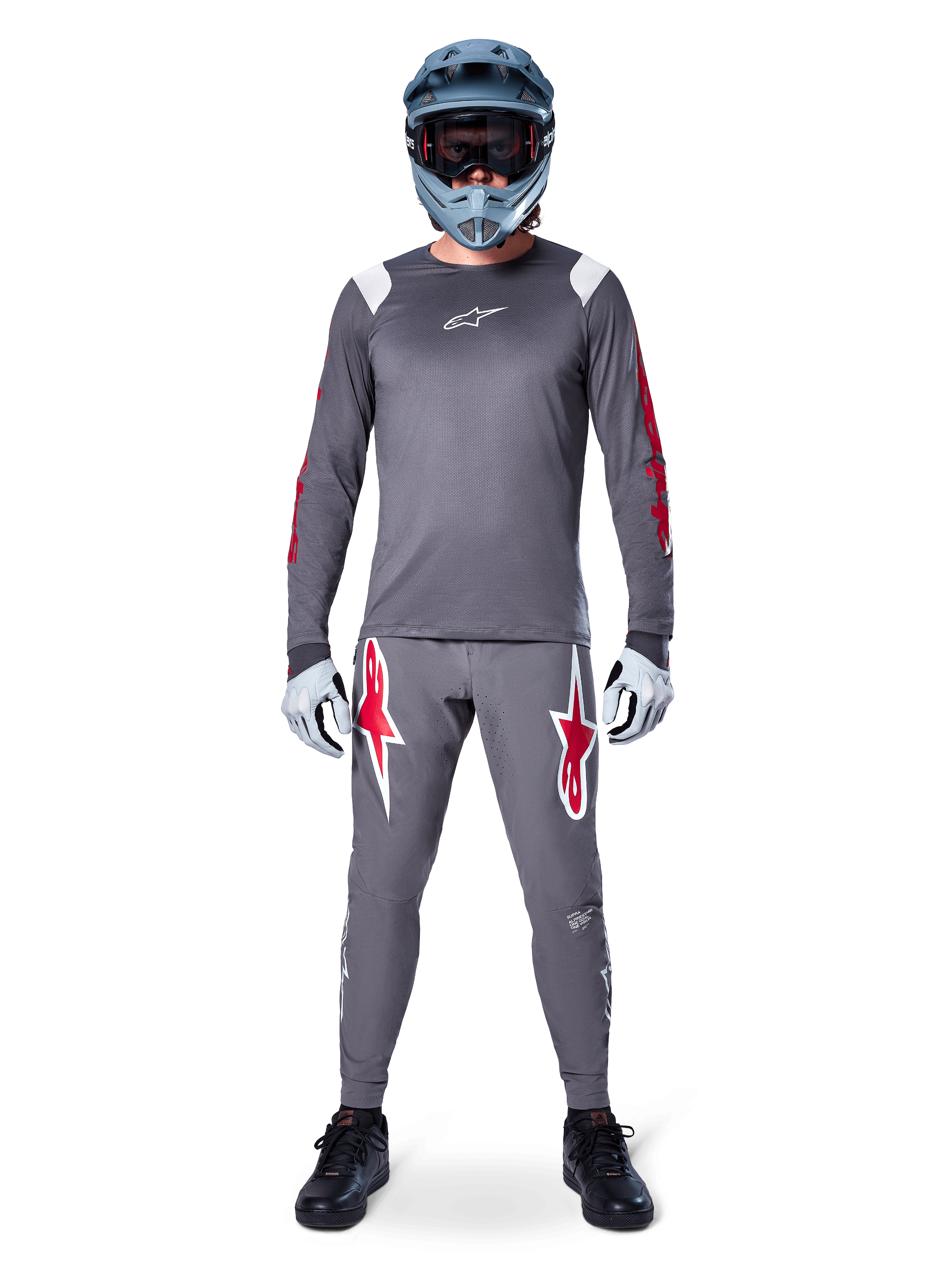 MTB rider wearing Alpinestars A-Supra Astar Long Sleeve Jersey, Dark Gray with red accents and white chest logo, matching gray pants with large red star graphics, blue full-face helmet with goggles, light gray gloves, and black shoes, professional mountain bike gear ensemble
