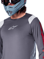 Alpinestars A-Supra Astar Long Sleeve Jersey, Dark Gray with white shoulder panels and red sleeve graphics, worn with a light blue-gray full-face helmet, performance mountain bike apparel