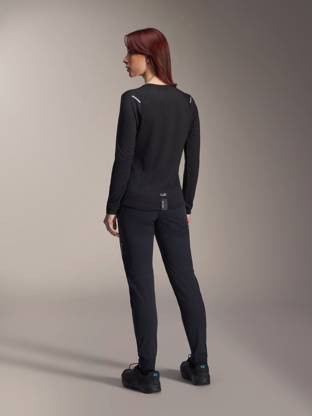 Alpinestars Woman Stella A-Dura Thermal Jersey, long-sleeve MTB jersey in Black, rear view showing reflective shoulder accents and extended drop tail, worn with black pants and shoes, performance gear for mountain biking.