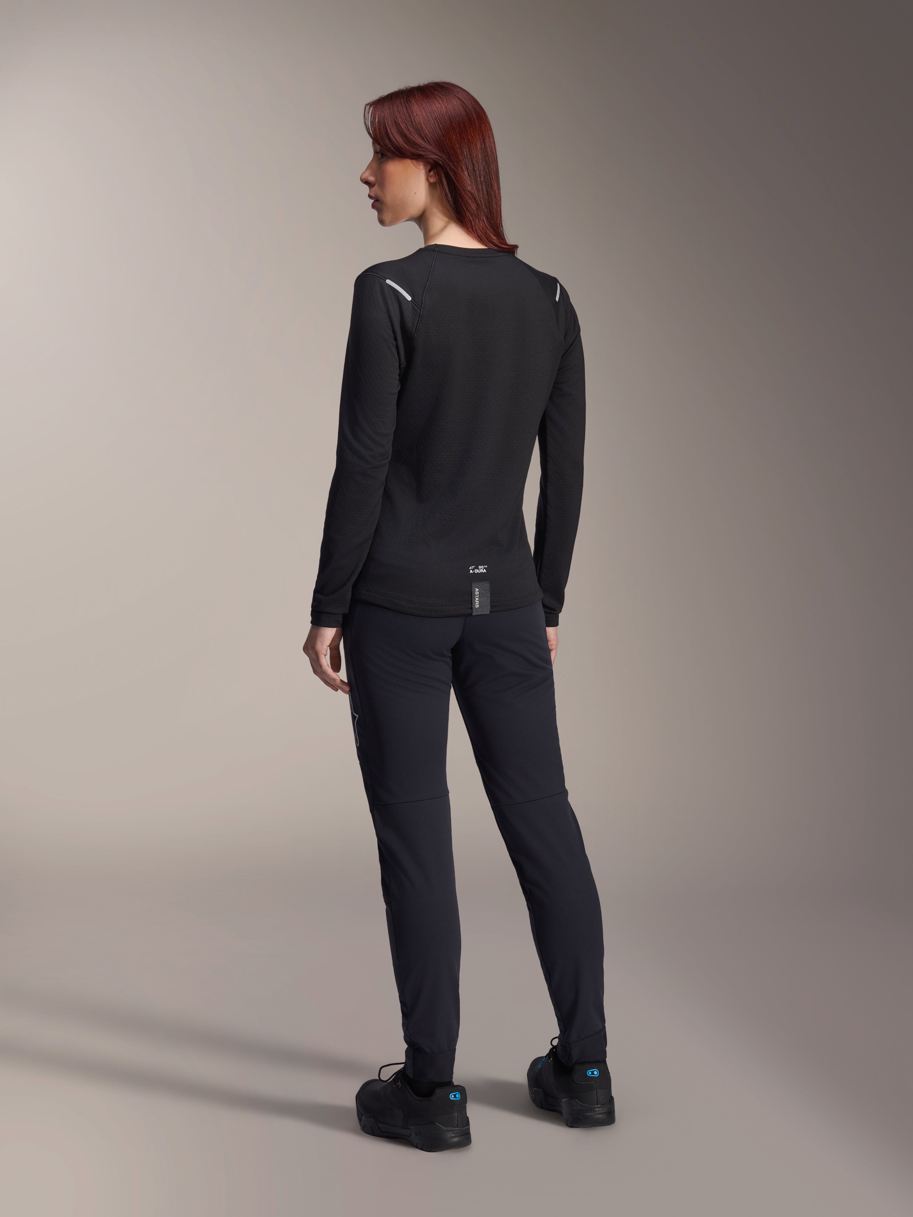 Alpinestars Woman Stella A-Dura Thermal Jersey, long-sleeve MTB jersey in Black, rear view showing reflective shoulder accents and extended drop tail, worn with black pants and shoes, performance gear for mountain biking.