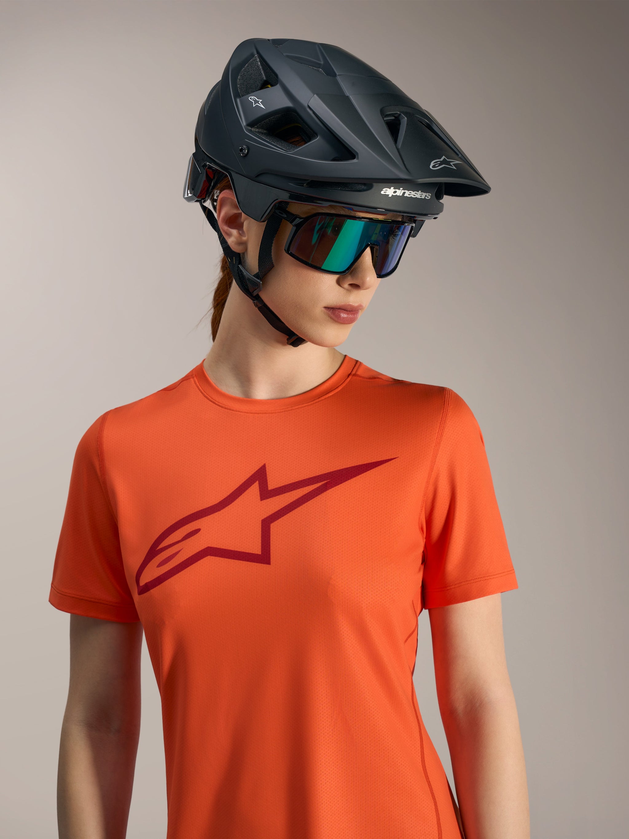 Orange Alpinestars Stella A-Dura Ride Short Sleeve Jersey with red logo, black MTB helmet, and iridescent sunglasses.