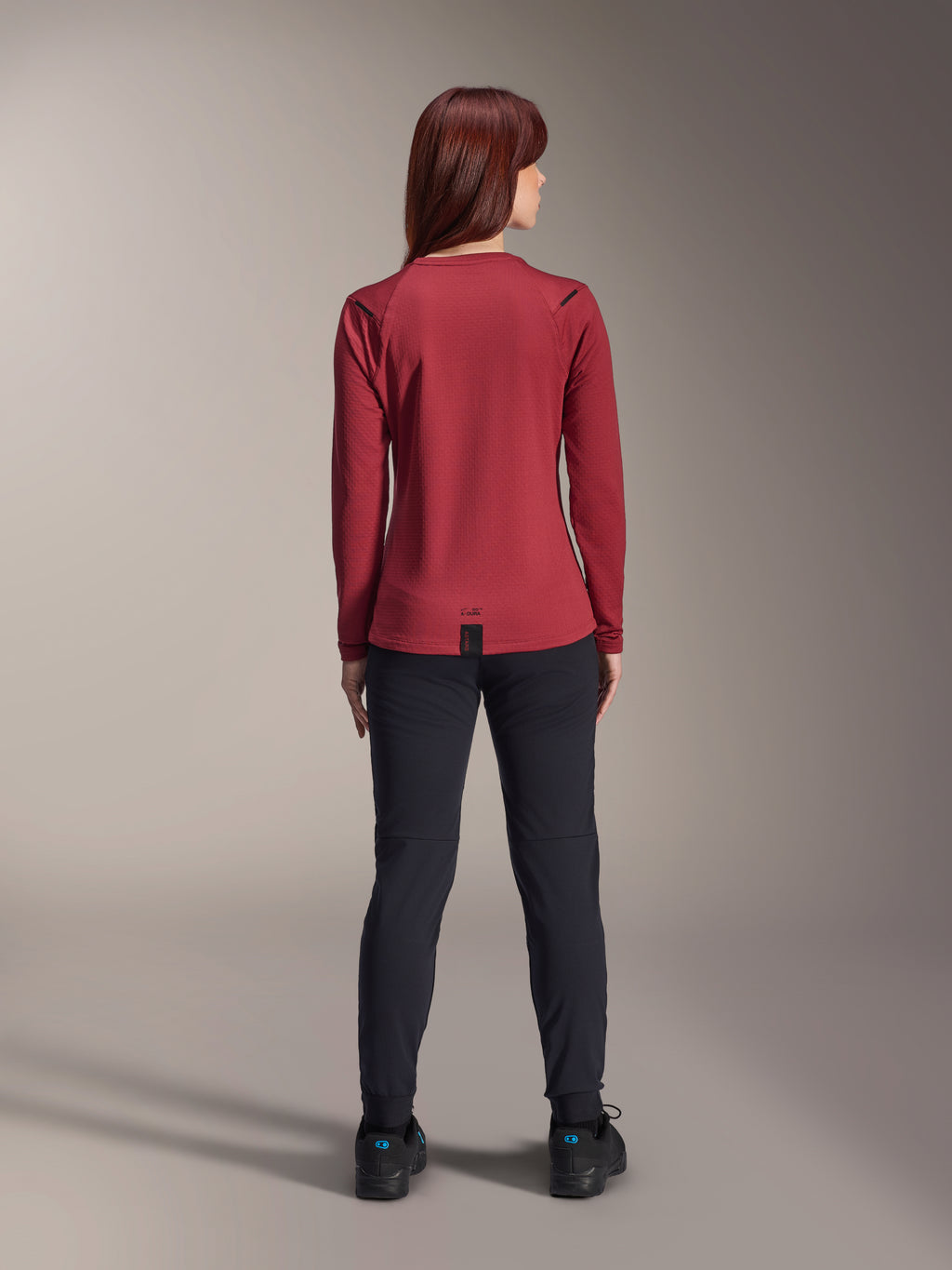 Woman wearing Alpinestars Stella A-Dura Thermal Jersey, Burgundy, rear view, long-sleeve insulated cycling jersey with an extended drop tail, paired with black pants and black riding shoes, outdoor mountain bike apparel.