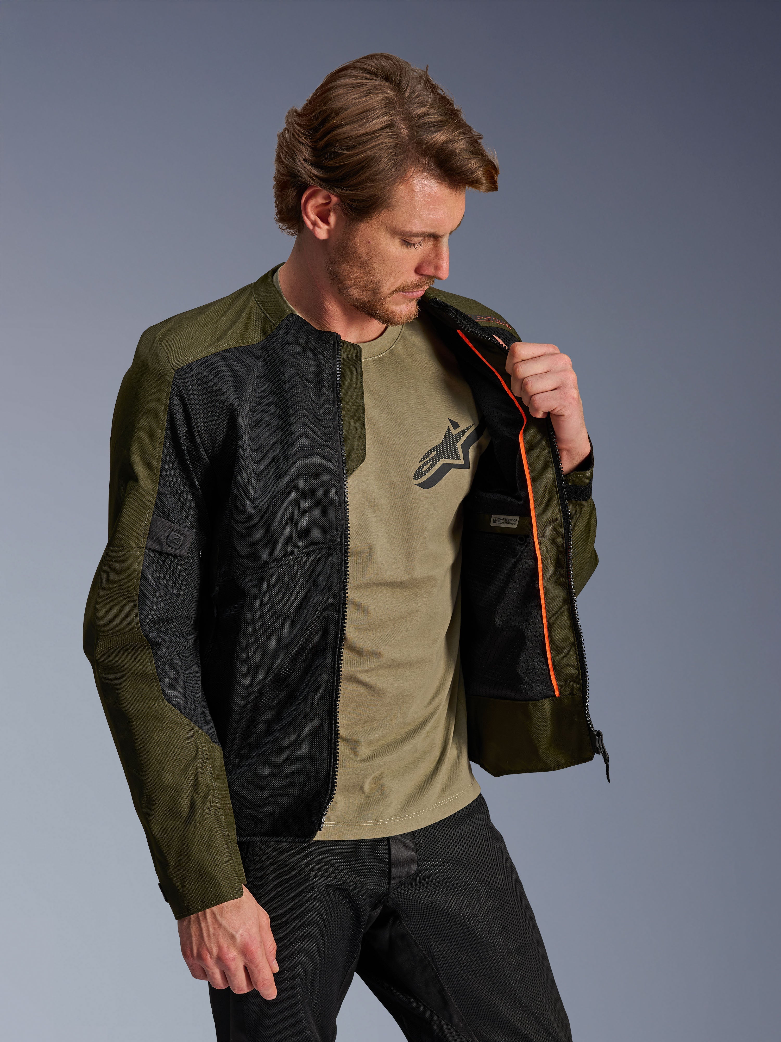 Alpinestars C-1 Air Jacket in Khaki and Black, worn by a male model to show the orange interior lining and waterproof compartment, featuring large tech mesh panels for airflow, urban contemporary motorcycle style paired with a matching olive t-shirt and black pants