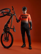 A-Dura ASTARS Long Sleeve Jersey 2.0 in orange-black gradient with bold chest logo, shown with full mountain bike gear.