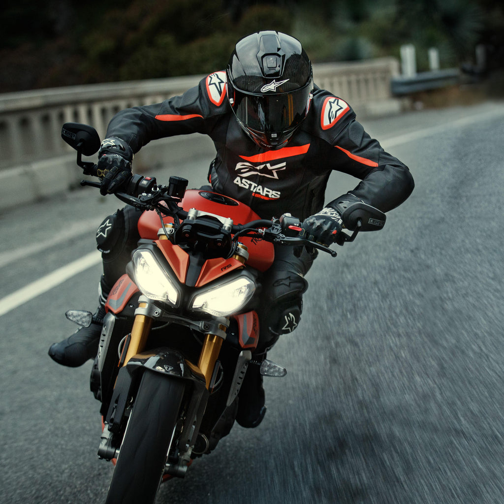 Motorcyclist riding a red sport bike, Alpinestars GP Plus R V4 Rideknit Leather Jacket, Black with Red Fluo and White details, featuring white Alpinestars logo and 'ASTARS' text, paired with black racing helmet and gloves, performance riding gear in action on a road.