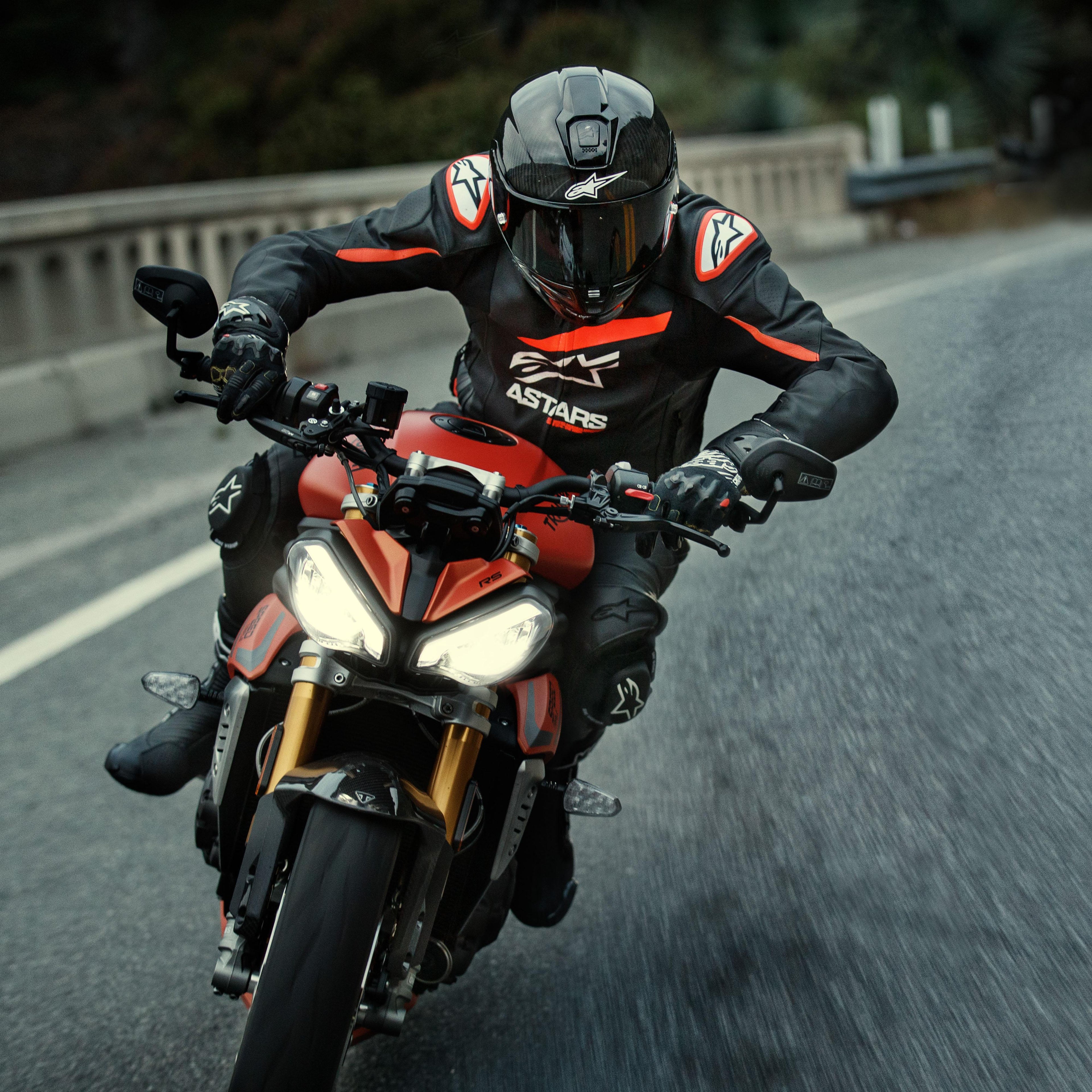Motorcyclist riding a red sport bike, Alpinestars GP Plus R V4 Rideknit Leather Jacket, Black with Red Fluo and White details, featuring white Alpinestars logo and 'ASTARS' text, paired with black racing helmet and gloves, performance riding gear in action on a road.