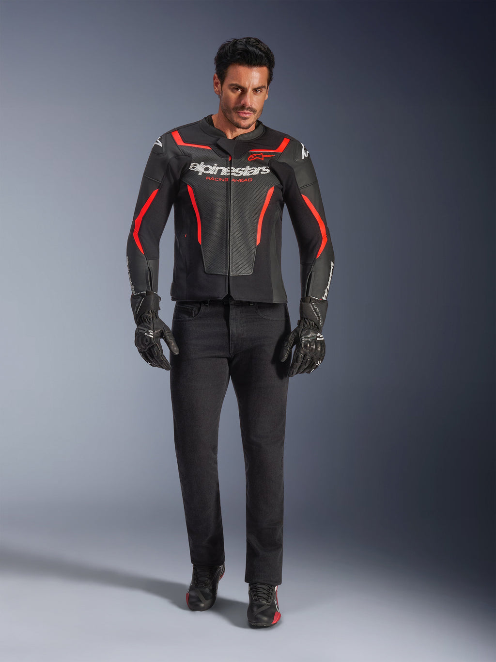 Male rider wearing Alpinestars GP Force V2 Air Leather Jacket, Black and Red Fluo, paired with black riding jeans, gloves, and shoes, featuring perforated leather construction, white logo branding, and sleek red accents for a sport-oriented look