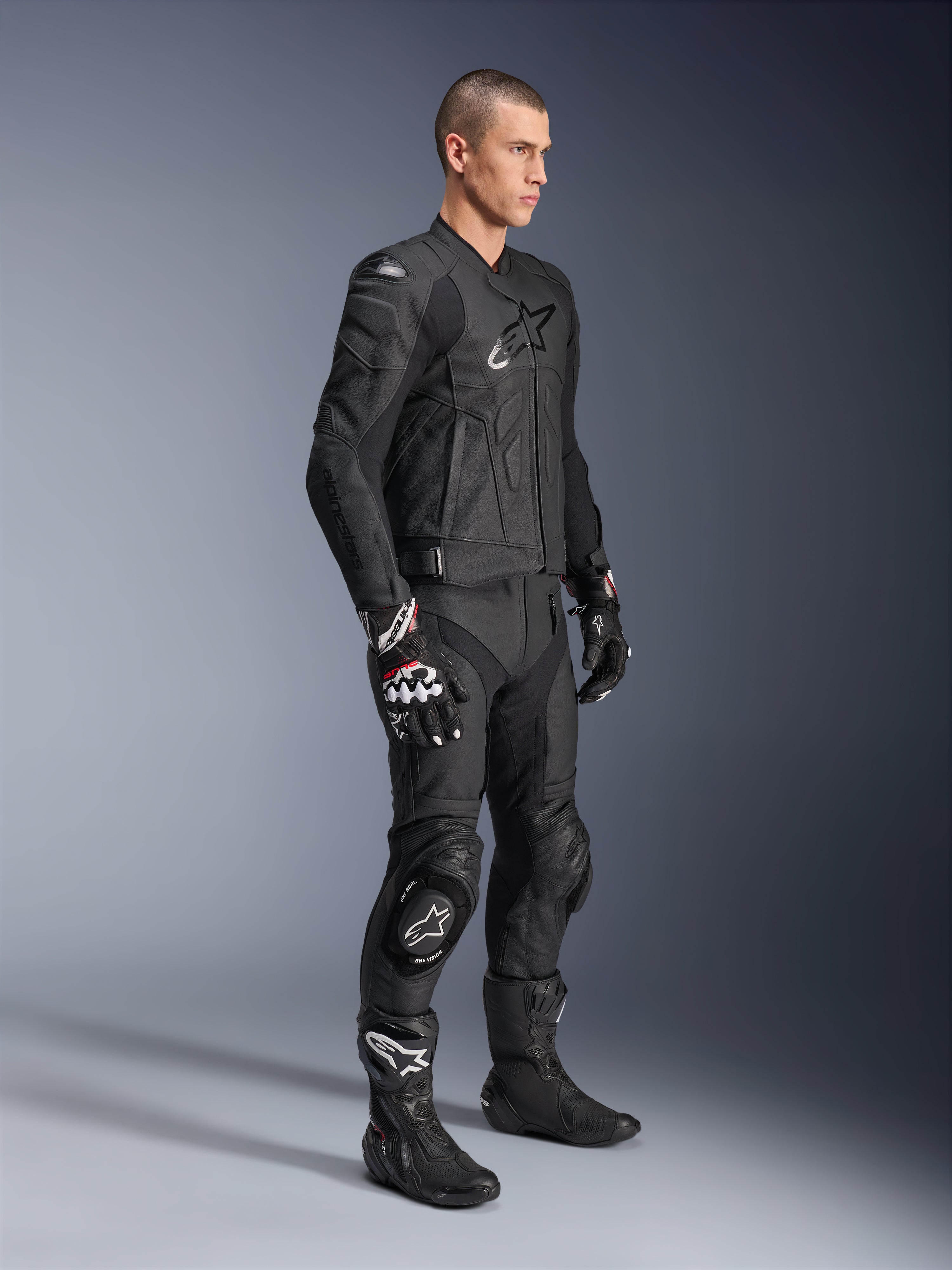 Alpinestars Dusk Leather Jacket, Black, worn by a male model with matching leather motorcycle pants, racing gloves, and boots, featuring a prominent star logo on the chest and ergonomic sport design for road riding