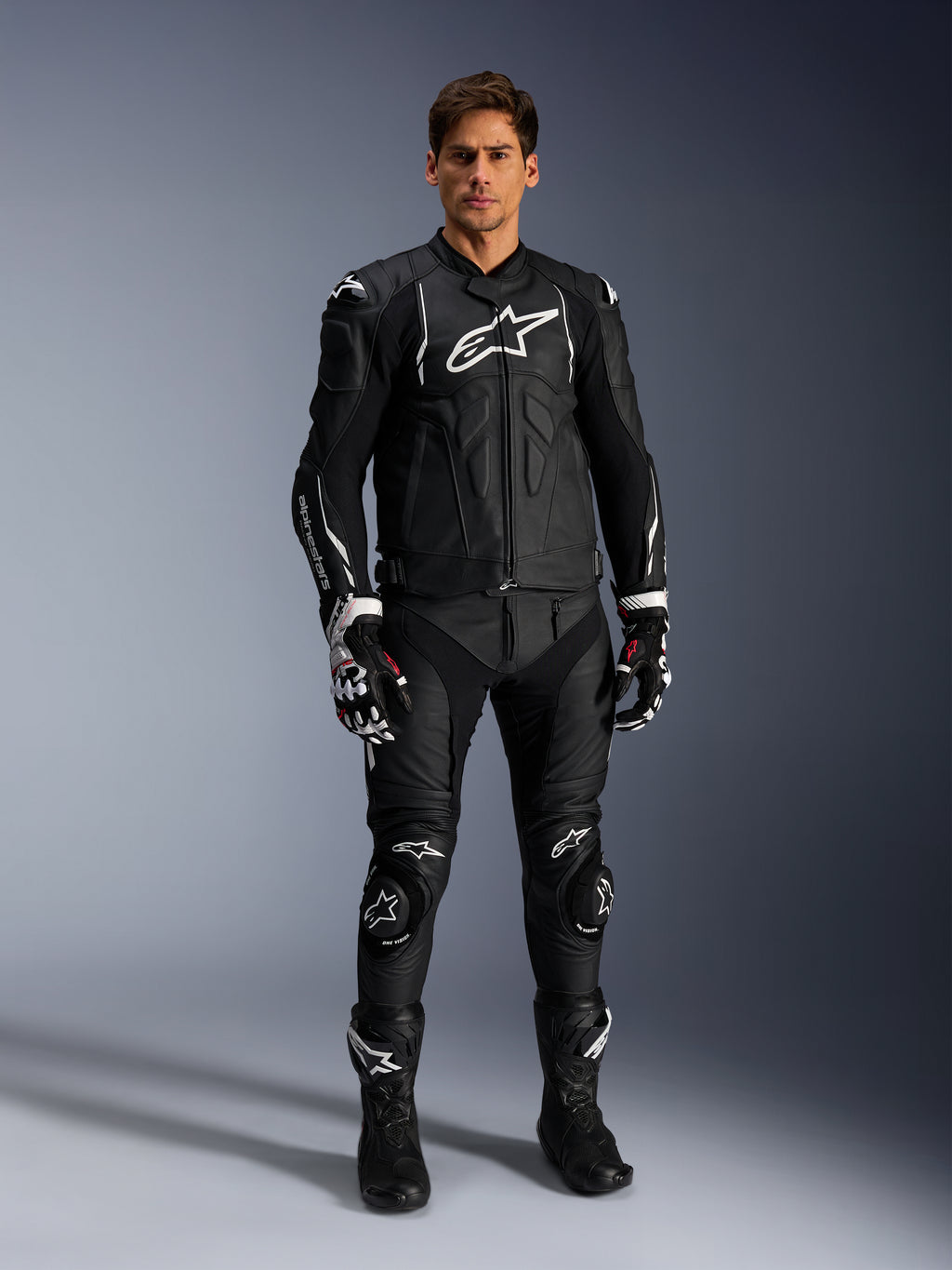 Male model wearing the Alpinestars Dusk Leather Jacket in Black and White, featuring prominent white logos on the chest and shoulders, paired with matching black leather motorcycle pants, black and white gloves, and black boots, complete sport racing outfit for road use.