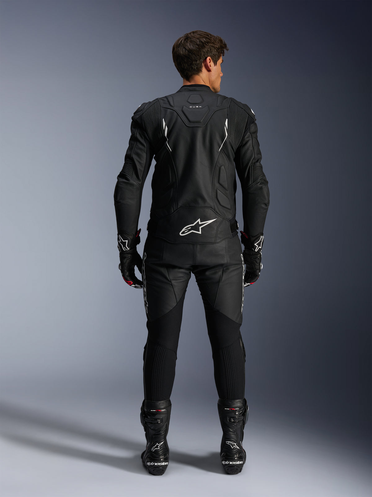 Motorcycle rider from a rear view wearing the Alpinestars Dusk Leather Jacket and Missile V3 Leather Pants, full black leather suit with white accents, jacket features 'Dusk' branding and Alpinestars logo on the back, paired with black racing boots and gloves, coordinated motorsport outfit for road racing.