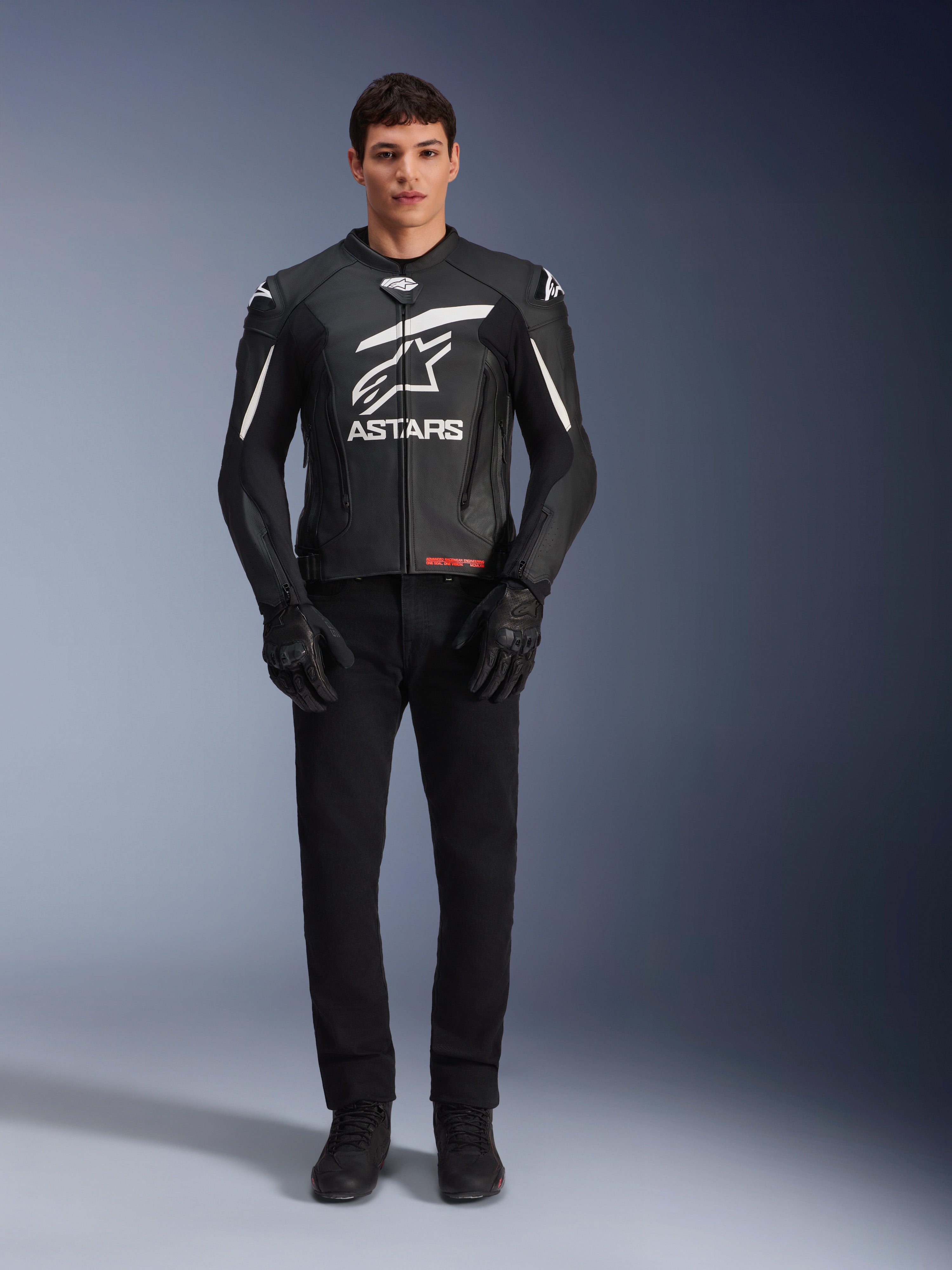 Male model wearing Alpinestars GP Plus V4 Leather Jacket, Black and White, featuring bold white Astars branding on the chest and white accents on the shoulders, paired with black riding gloves and pants, sleek sport-oriented motorcycle apparel