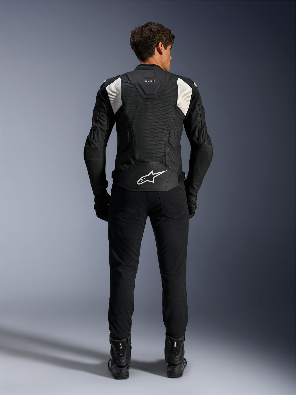 Alpinestars Dusk Airflow Leather Jacket, Black and White, rear view of a rider wearing black pants and boots, featuring perforated leather for ventilation, white accordion stretch panels on the shoulders, and white branding on the lower back