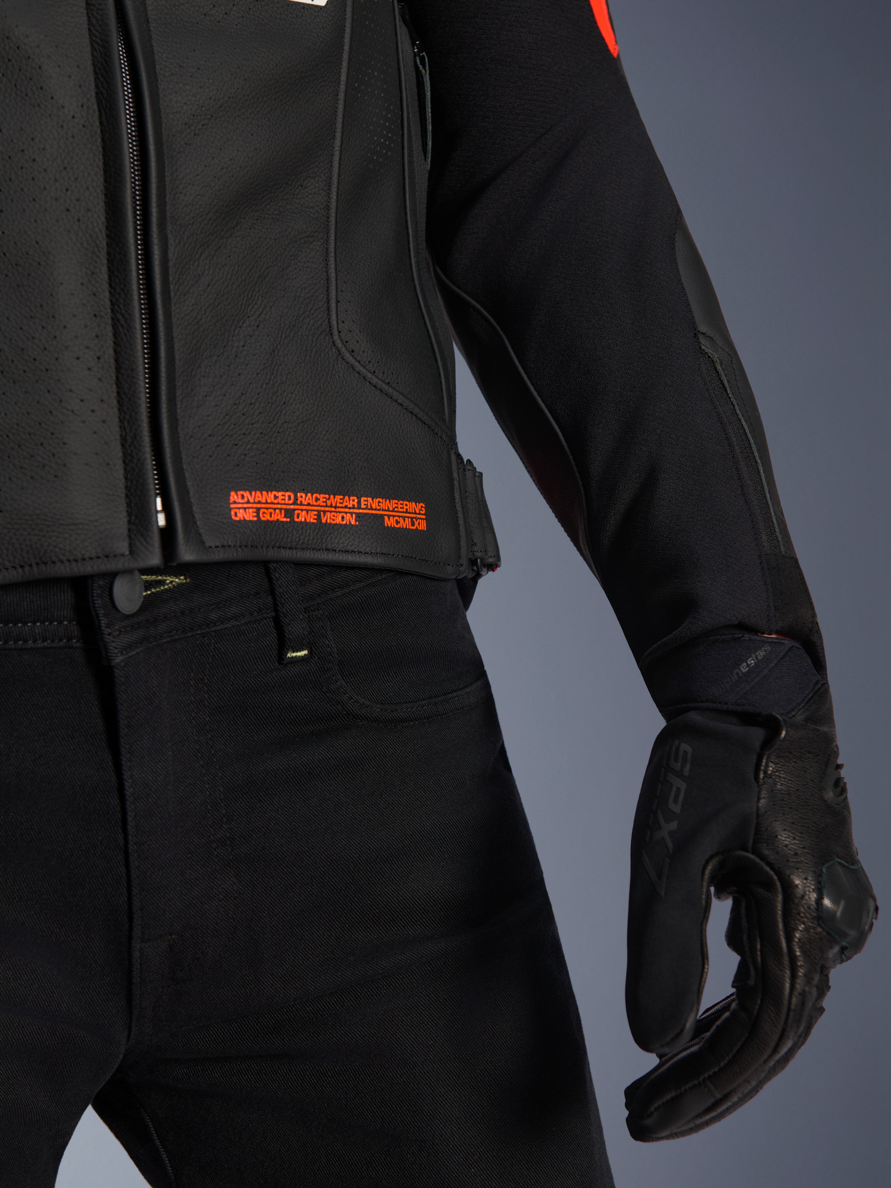 Alpinestars GP Plus R V4 Airflow Leather Jacket, close-up of waist and sleeve, black perforated leather with red fluo text branding reading 'Advanced Racewear Engineering', paired with black riding pants and SPX 7 motorcycle glove