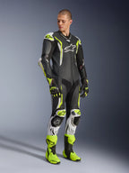 Alpinestars GP Tech V5 One Piece Leather Suit, Black White and Yellow Fluo, worn by a model with matching racing gloves and boots, professional motorcycle racing gear featuring aerodynamic performance and protective armor.
