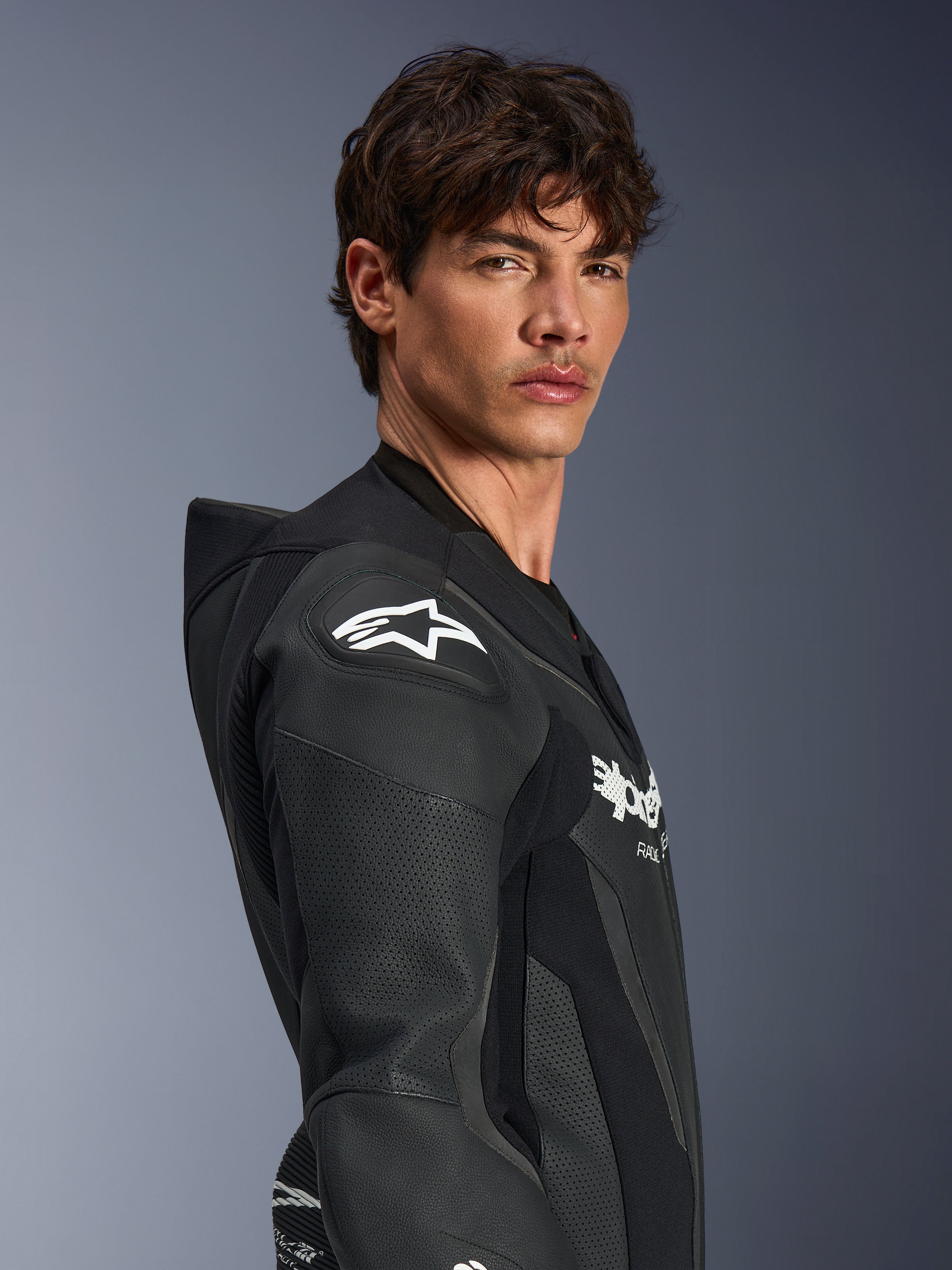 Male model wearing Alpinestars GP Force V2 Leather Suit 1-Piece in Black, side profile view highlighting the white Alpinestars star logo on the DFS Lite shoulder protector, perforated leather panels, and aerodynamic speed hump.