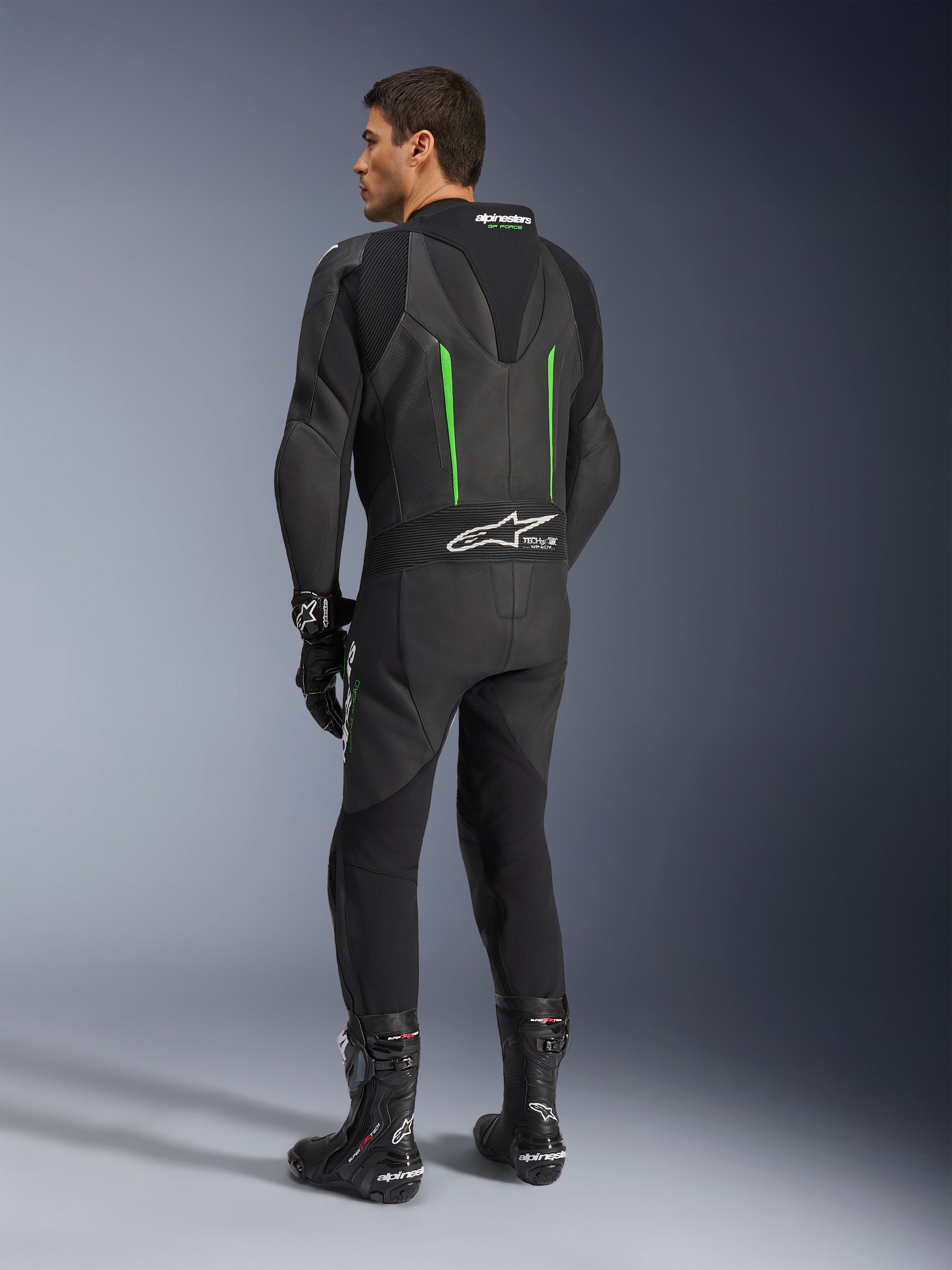 Alpinestars GP Force V2 Leather Suit, 1-Piece Motorcycle Suit, Black with Green accents, back view showing the aerodynamic speed hump and white logo on the accordion stretch panel, paired with black racing boots and gloves.