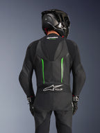 Rear view of a rider wearing the Alpinestars GP Force V2 Leather Suit in Black Green, featuring an aerodynamic speed hump, Tech-Air Ready branding on the lower back accordion panel, and perforated leather construction, paired with a black carbon fiber racing helmet.